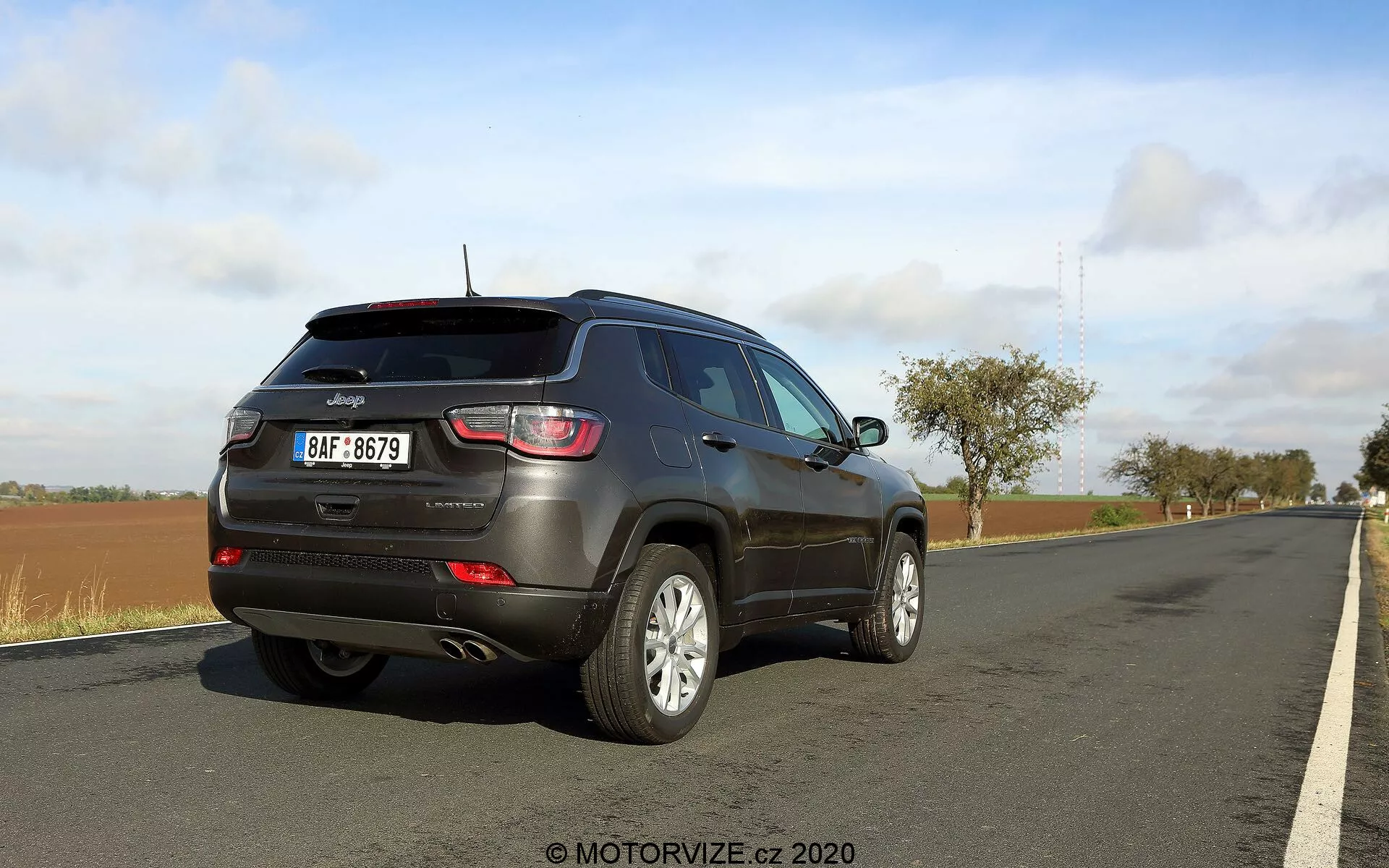 Vista di tre quarti posteriori di una Jeep Compass 2017. L'immagine evidenzia il profilo laterale e il retro della vettura, mostrando luci posteriori distintive, un tergicristallo posteriore, uno spoiler sul tetto, riflettori sul paraurti posteriore e ruote in lega a più razze. Il veicolo è parcheggiato sul ciglio di una strada con un bordo erboso accanto. Il cielo è parzialmente nuvoloso, indicando condizioni meteorologiche variabili.