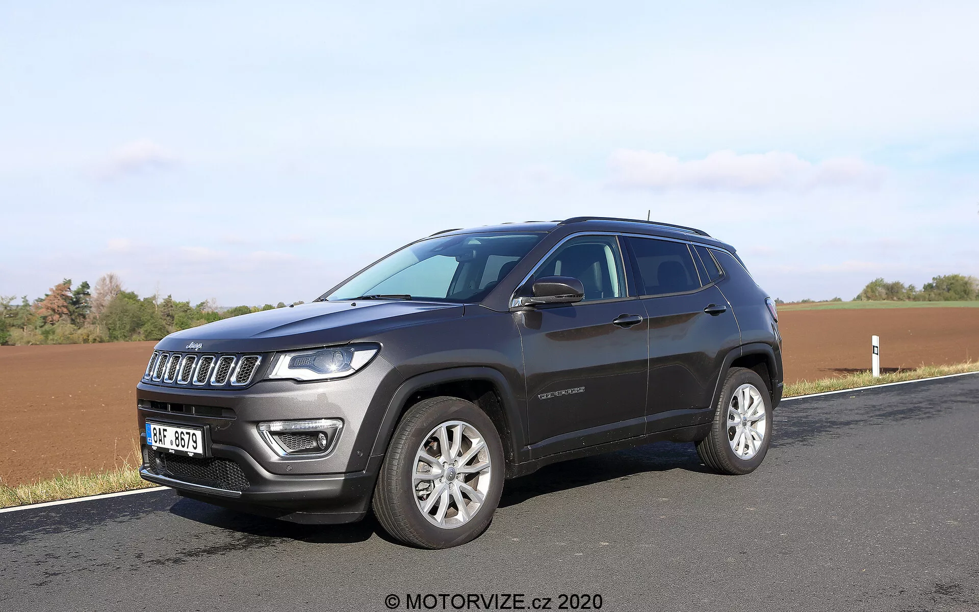 SUV moderno parcheggiato lungo una strada con un campo sullo sfondo, catturato da un angolo frontale di tre quarti. La foto mette in evidenza la griglia frontale a sette slot del SUV, i fari distintivi dotati di LED, i cerchi in lega e la vernice metallica scura. Sono visibili dettagli laterali come la linea dei finestrini, le maniglie delle porte, gli specchietti laterali, possibilmente con indicatori di direzione, e una linea del tetto leggermente inclinata. È visibile anche un badge sulla porta anteriore, che fornisce potenziali informazioni sul livello di allestimento.