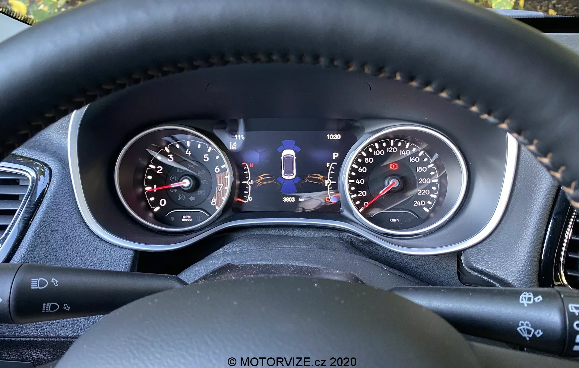 Vista ravvicinata del quadro strumenti del Jeep Compass 2017 dalla prospettiva del conducente, che mostra sia indicatori analogici che un display digitale centrale. Caratteristiche includono un contagiri, tachimetro a doppia unità, indicatore di selezione del cambio, indicatore del carburante, contachilometri e ulteriori indicatori di tecnologie di sicurezza come l'assistenza al mantenimento della corsia. La cucitura sul volante in materiale simile alla pelle evidenzia materiali di qualità e comfort, mentre le leve di controllo e i pulsanti suggeriscono caratteristiche avanzate di convenienza e assistenza al conducente.