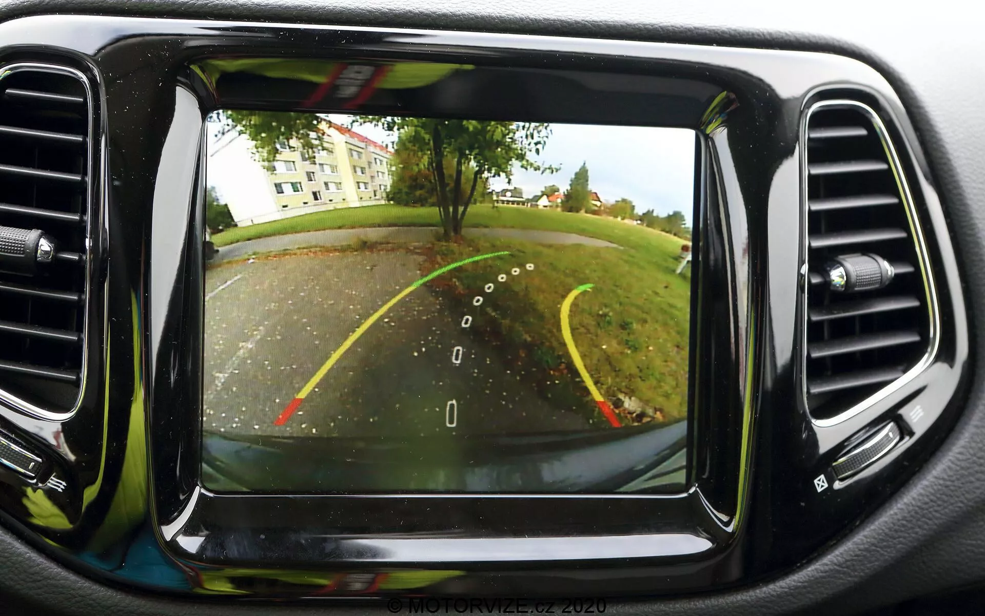 Immagine del sistema di telecamera di retromarcia sul display del cruscotto di una Jeep Compass 2017. La telecamera, posizionata sul retro vicino alla targa, fornisce una vista grandangolare dietro il veicolo. Sono visibili linee guida per la retromarcia, che indicano la larghezza dell'auto e il percorso previsto con linee gialle e rosse per la distanza. La scena mostra un parcheggio o un vialetto con alberi e area erbosa sullo sfondo. Lo schermo è posizionato centralmente sul cruscotto, circondato da prese d'aria per il condizionamento, e presenta una qualità dell'immagine chiara, tipica delle tecnologie automobilistiche moderne.