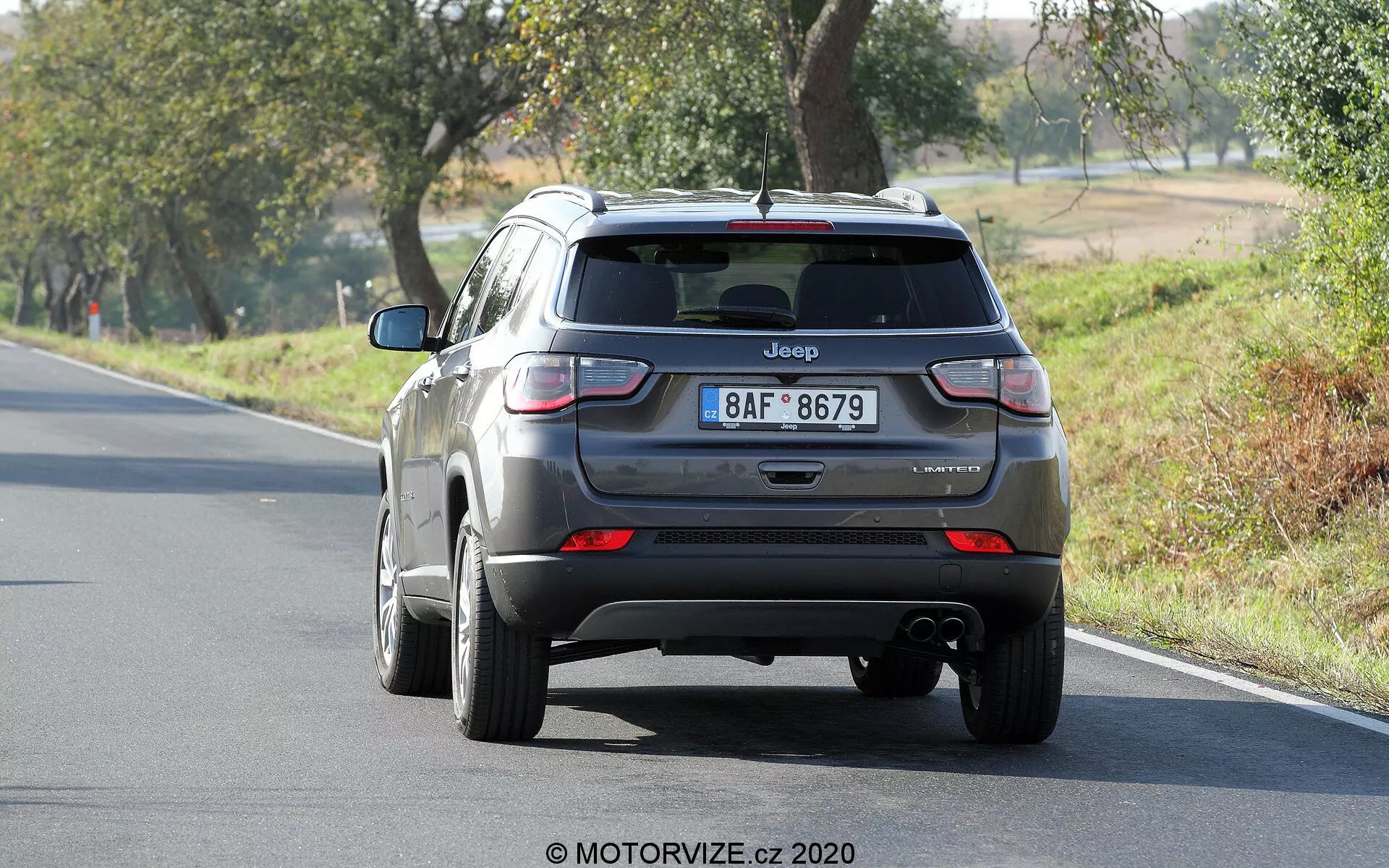 Vista posteriore di una Jeep Compass 2017 argento che mostra il portellone, il paraurti posteriore e le luci posteriori. È visibile la scritta 'Jeep' e il nome del modello 'Compass', con il badge del livello di allestimento leggermente poco chiaro. L'immagine mostra chiaramente le luci posteriori, il terminale di scarico sotto il paraurti e l'auto posizionata su una strada a due corsie circondata da alberi in un ambiente rurale.
