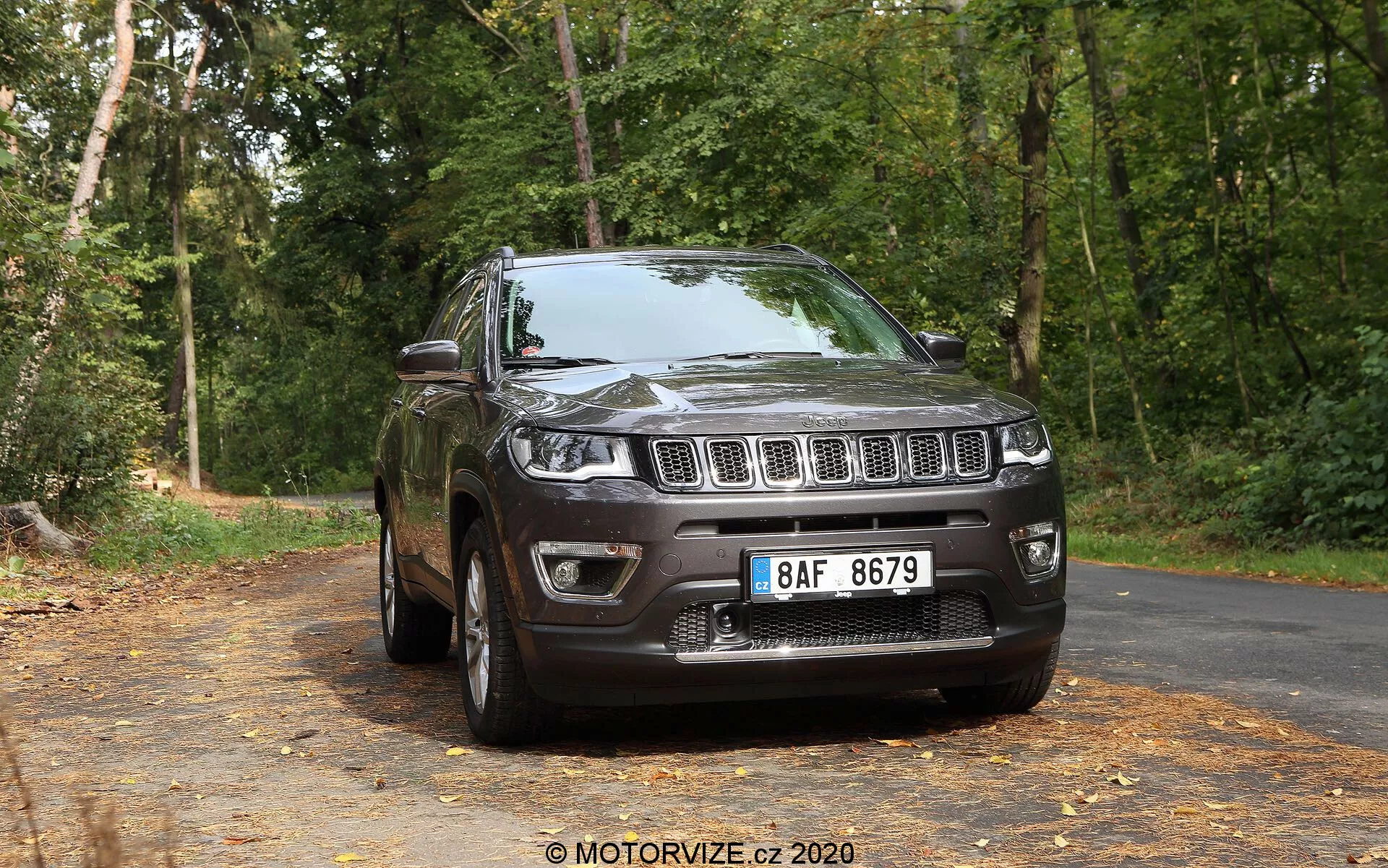 Vista tre quarti anteriore di un Jeep Compass SUV del 2017 di colore scuro su una strada asfaltata ricoperta di foglie, con un fondale di alberi in un ambiente boschivo. L'immagine mostra la griglia anteriore con fessure verticali distintive, affiancata da fari che si estendono nei parafanghi, un paraurti anteriore ben definito con fendinebbia e un cofano rialzato e scolpito. Il profilo laterale rivela cerchi in lega, un parafango visibile, specchietti laterali, maniglie delle porte, contorni dei finestrini e un sistema di binari sul tetto, il tutto illuminato da luce naturale.
