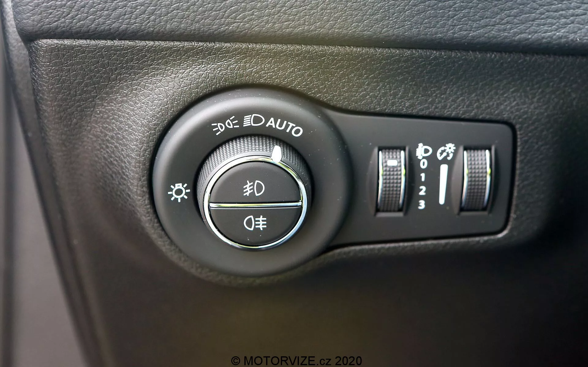 Vista ravvicinata dell'interruttore di controllo dei fari della Jeep Compass 2017, situato sul cruscotto a sinistra del volante. L'immagine, scattata con un leggero angolo, mette in evidenza la manopola rotante con impostazioni per luci di posizione, luci diurne, fari automatici, abbaglianti, anabbaglianti e posizione OFF. Controlli aggiuntivi a destra includono un cursore verticale per la luminosità del cruscotto e un pulsante che potrebbe essere per abbaglianti automatici o livellamento fari. L'interruttore è montato su una superficie di plastica nera testurizzata, con chiari simboli bianchi e riflessi leggermente lucidi.
