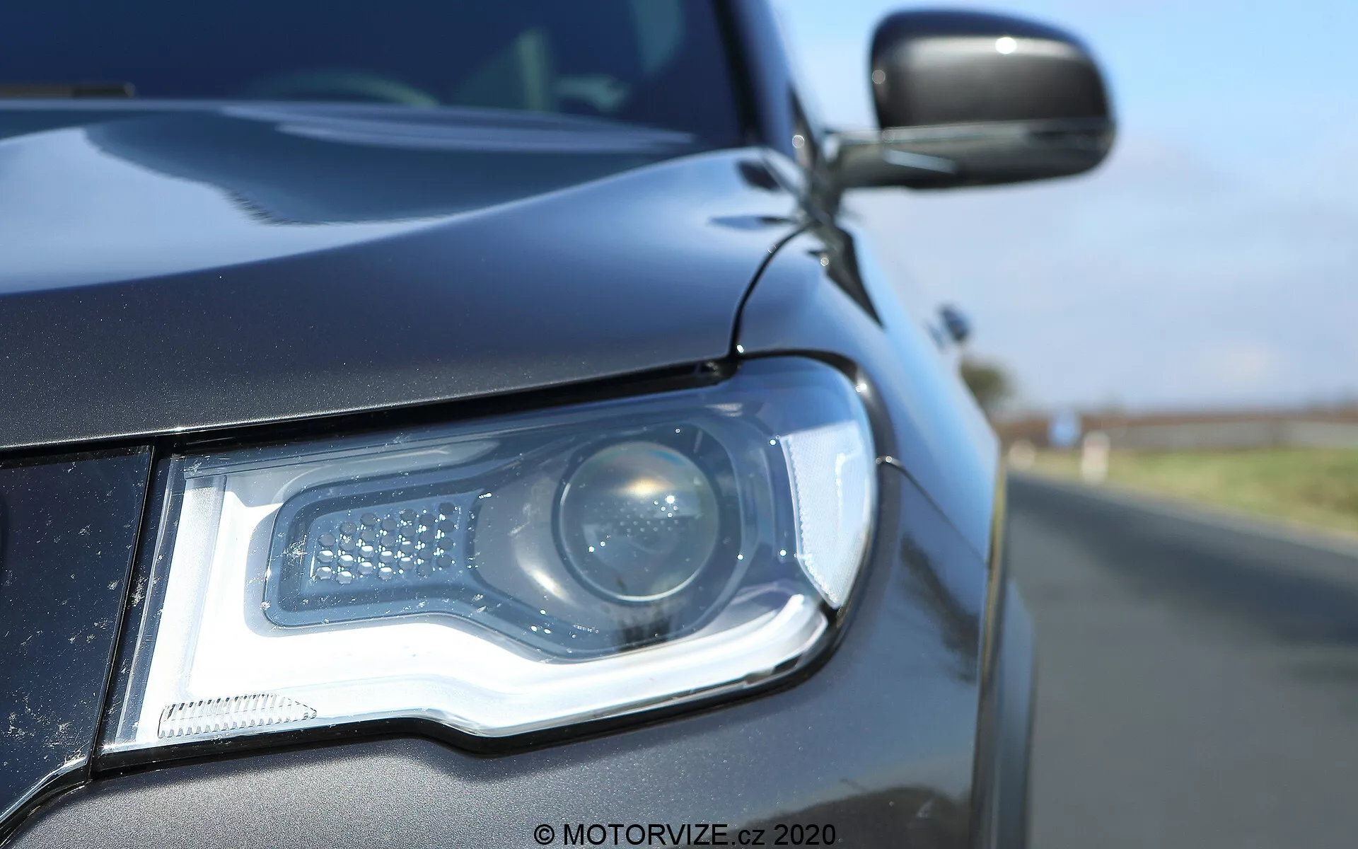 Vista ravvicinata del lato anteriore sinistro di una Jeep Compass 2017, che mostra l'assemblaggio dei fari da un'angolazione bassa a livello del suolo. Il moderno faro dispone di un fascio proiettore e luci diurne a LED disposte in un motivo distintivo. L'alloggiamento del faro, elegante, si integra perfettamente con la carrozzeria di colore scuro, riflettendo un cielo blu chiaro. Lo specchio laterale, parzialmente visibile, sembra abbinarsi al colore della carrozzeria e potrebbe includere un indicatore laterale. La scarsa profondità di campo sottolinea i dettagli del faro e della griglia anteriore contro uno sfondo sfocato della strada.