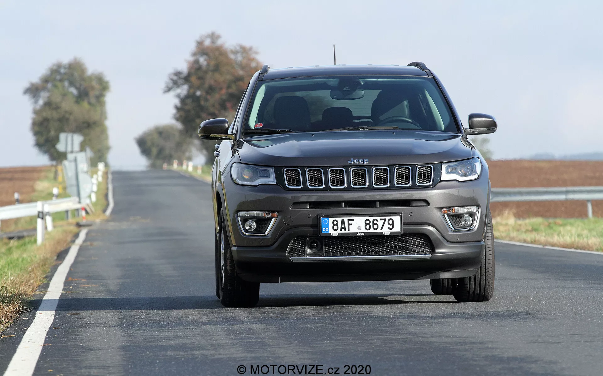 Vista frontale della Jeep Compass 2017 in movimento su una strada a due corsie, ripresa da un'angolazione bassa, con evidenziata la griglia prominente a sette slot, i fari quadrati con luci di marcia diurna moderne e i fendinebbia visibili con sensori di parcheggio anteriori sul paraurti. La strada si estende fino all'orizzonte con una doppia linea gialla continua, indicando una zona di divieto di sorpasso. Lo sfondo mosso indica che il veicolo è in movimento, mostrando l'assetto rialzato tipico dei SUV.