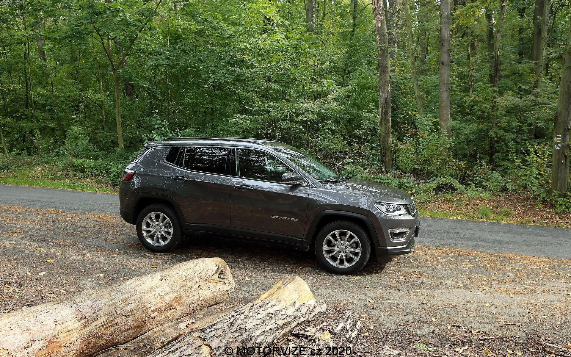 Vista laterale di un SUV Jeep Compass 2017 parcheggiato su una strada asfaltata in un ambiente naturale con lussureggianti alberi verdi. Il veicolo mostra linee di design moderne lungo le porte e i passaruota, con cerchi in lega visibili dal design multirazza. Ha una alta altezza da terra che suggerisce capacità fuoristrada. L'immagine cattura parte della griglia anteriore e dei fari, indicando uno stile contemporaneo. Sono presenti portapacchi, indicatori di direzione integrati negli specchietti laterali e finestre oscurate. La vernice di colore scuro con finitura lucida indica un veicolo ben mantenuto. Mentre è fermo, il SUV è mostrato con abbondante luce naturale, ideale per mostrare le sue caratteristiche e l'attrattiva per gli appassionati di attività all'aperto e avventura.