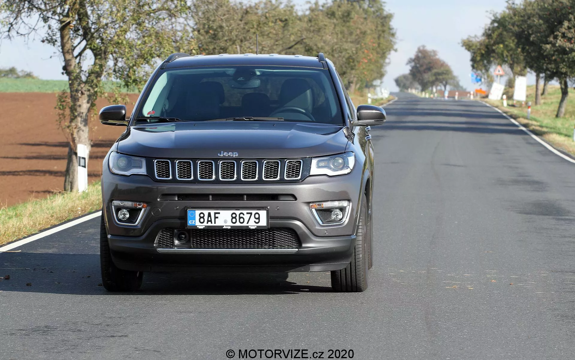 Vista frontale di una Jeep Compass 2017 che viaggia su una strada rurale o suburbana a due corsie, catturata da un angolo basso, mettendo in mostra la sua iconica griglia a sette slot, DRL LED, fari fendinebbia e i contorni robusti del cofano. L'auto è centrata e relativamente vicina alla fotocamera, con un lieve sfocatura delle ruote e dello sfondo. Il cielo sereno suggerisce una giornata luminosa e soleggiata.