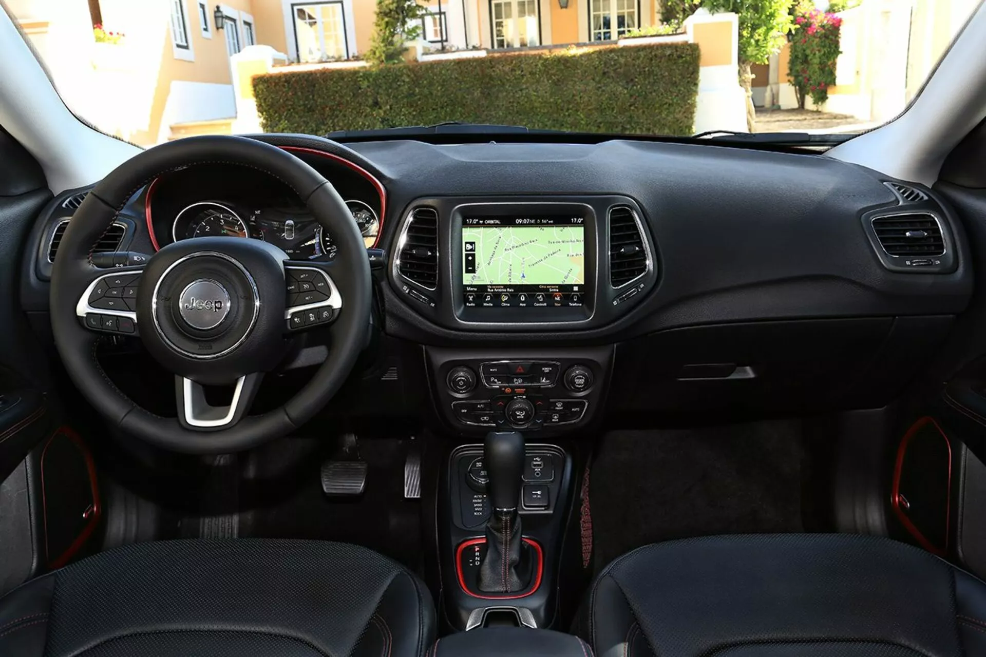 Vista interna del Jeep Compass 2017 dalla prospettiva del guidatore che mostra il volante con comandi integrati, cruscotto, console centrale con sistema di navigazione, controlli climatici, leva del cambio automatica con dettagli rossi sportivi, pulsante di avvio-stop senza chiave e porta per la connettività multimediale