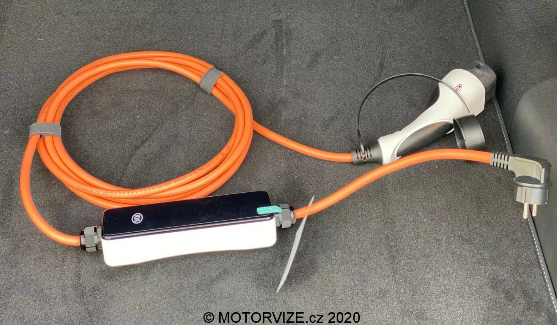 Cavo di ricarica e adattatore per veicolo elettrico (EV) del Kia Ceed 2018, con un EVSE (Apparecchiatura di Alimentazione del Veicolo Elettrico) portatile arancione ad alta visibilità per la sicurezza. La configurazione include un connettore e una spina di ricarica per l'uso in presa domestica, nonché una scatola di controllo in linea con indicatori LED, tutto posizionato sulla superficie tappezzata del bagagliaio o dell'area di carico del veicolo.