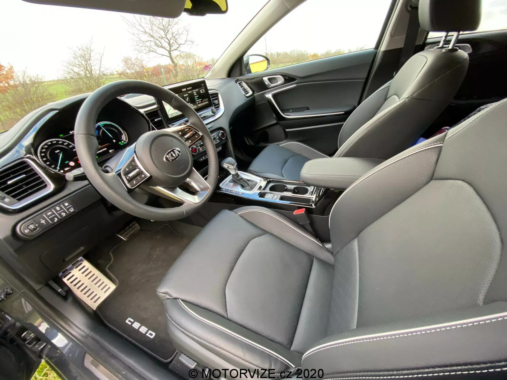 Interno di una Kia Ceed 2018 visto dalla prospettiva del passeggero, catturando l'area anteriore con attenzione al cruscotto, console centrale e sedile del conducente. È visibile il volante con più pulsanti di controllo e il logo Kia, insieme a un quadro strumenti digitale parzialmente visibile. Al centro del cruscotto si trova un grande schermo touchscreen di infotainment circondato da comandi manuali per il climatizzatore. La console centrale include una leva del cambio, portabicchieri e manopole di controllo. Sono altresì visibili il sedile anteriore rivestito in pelle e il pannello della porta di materiali misti, suggerendo un allestimento di lusso. Anche i pedali freno e acceleratore in metallo indicano un tocco sportivo premium.