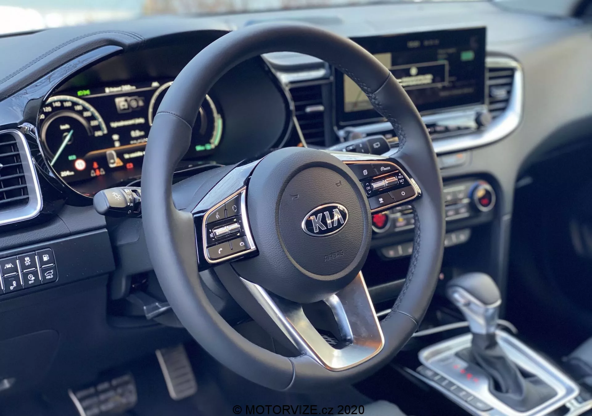 Vista in prima persona degli interni della Kia Ceed 2018 che evidenzia il volante multifunzione a tre razze moderno con logo Kia e quadro strumenti digitale, che mostra il tachimetro e il possibile contagiri, insieme al sistema di infotainment touchscreen, ai controlli del clima, alla leva del cambio automatico color argento e alla dettagliata consolle centrale.