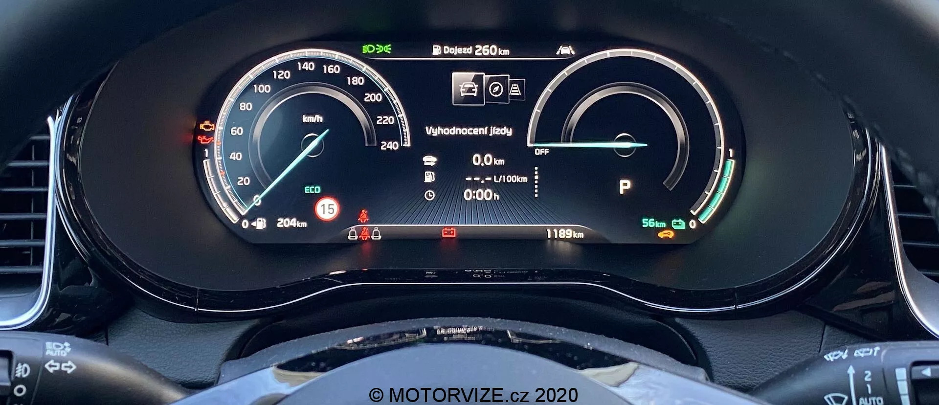 Vista ravvicinata del cruscotto digitale della Kia Ceed 2018 dal sedile del guidatore. Il display include un tachimetro con ago fisico e lettura digitale che mostra 15 km/h. A destra, c'è un indicatore di potenza/batteria che indica una batteria completamente carica con un'autonomia di 481 km. Altri elementi includono il display dello stato pronto, l'informazione del conducente con luci di avviso, una lettura della temperatura esterna di 20,5°C e il veicolo in modalità Park. È visibile l'arco superiore del volante, con un design sportivo e fondo piatto.