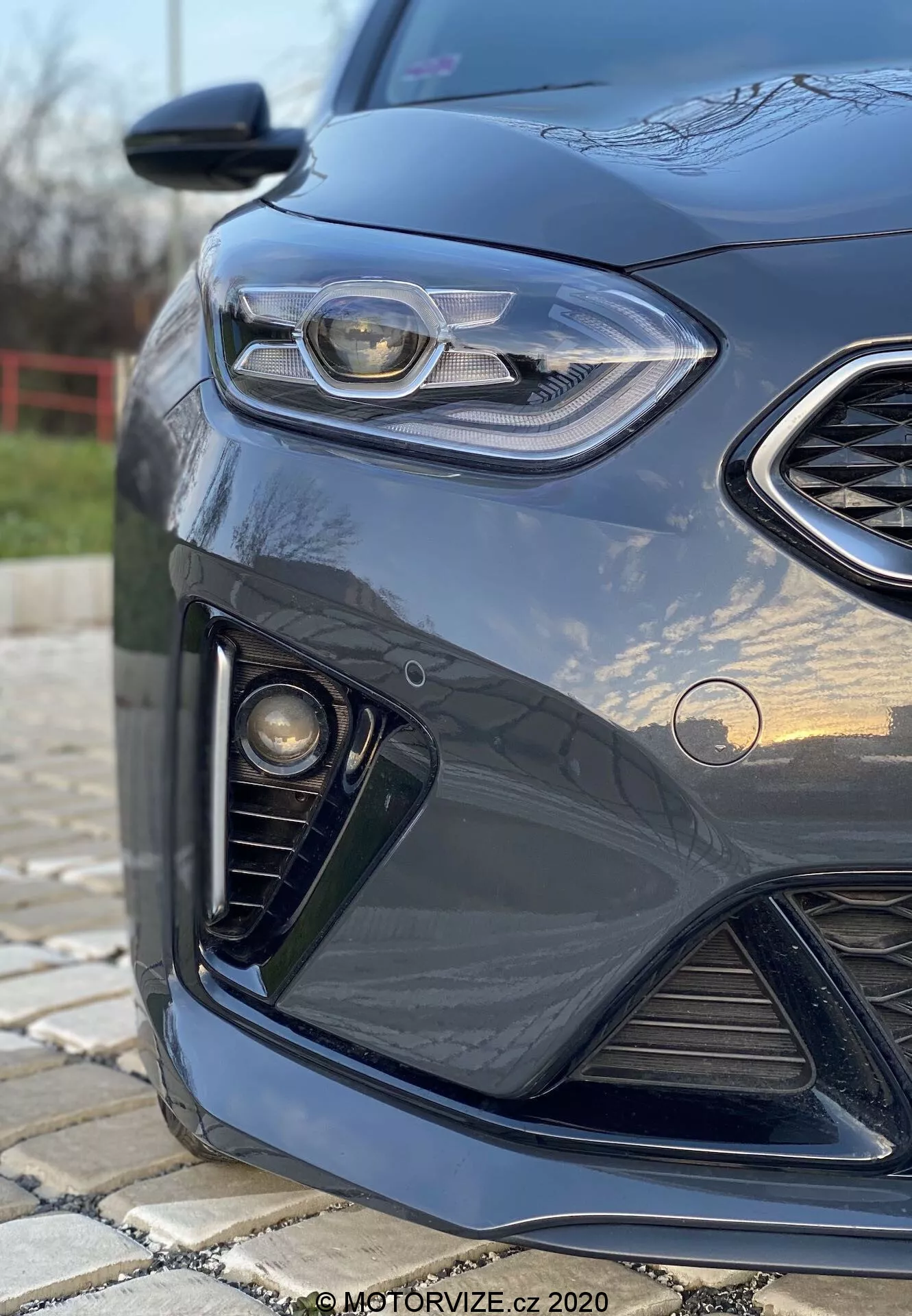 Vista angolata ravvicinata del lato anteriore sinistro di una Kia Ceed 2018 scura, con evidenza del design elegante dei fari con luce diurna a LED, griglia a nido d'ape, gruppo di luci fendinebbia e paraurti anteriore aerodinamico, con sfondo sfocato e riflessi sulla vernice lucida.