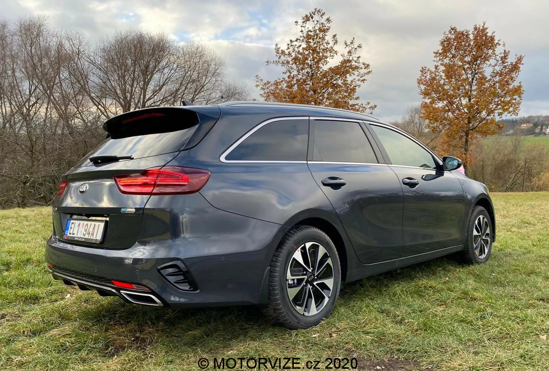 Vista posteriore tre quarti di un'auto moderna hatchback o station wagon, con finitura grigio scuro, fanali posteriori strutturati verticalmente, sistema di scarico doppio e ruote in lega a più razze, parcheggiata su un'area erbosa con una leggera inclinazione, circondata da alberi e un cielo coperto.