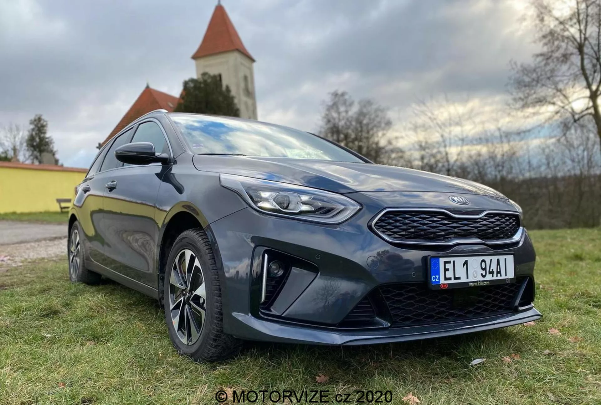 Vista dettagliata di una moderna Kia Ceed 2018 hatchback fotografata da un angolo basso di tre quarti frontale, che mostra la griglia prominente, i fari a LED angolari e il profilo laterale fino alla porta del conducente. L'auto è di colore scuro, probabilmente blu scuro o nero, con una lucentezza lucida che evidenzia il suo stato ben mantenuto. I principali elementi di design includono una griglia frontale con motivo a pattern, fari affilati con lenti chiare, elementi aerodinamici pronunciati sul paraurti anteriore inferiore, cerchi in lega multirazza, specchi laterali color carrozzeria con indicatori integrati e una gonna laterale sportiva. La postura dell'auto suggerisce un equilibrio tra comfort e sportività. La luce naturale del giorno con illuminazione soffusa esalta i dettagli e sullo sfondo si vede un paesaggio naturale con una chiesa o una torre.