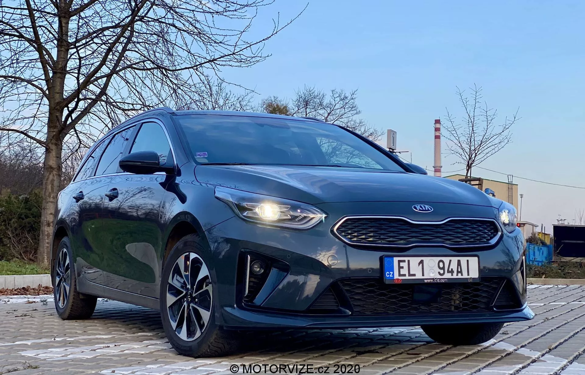 Fotografia della Kia Ceed 2018 scattata da un'angolazione bassa e anteriore sinistra, che enfatizza la griglia, i fari e il design della parte anteriore del veicolo. Lo stile aggressivo dell'auto è messo in evidenza dai fari prominenti, dalla griglia a naso di tigre e dai contorni del cofano. L'immagine mostra anche le luci diurne a LED, il paraurti anteriore aerodinamico e i cerchi in lega dal design elegante. L'ambiente è all'aperto con alberi e strutture industriali sullo sfondo, creando un contrasto. La vernice metallizzata scura del veicolo è lucida e ben illuminata, con riflessi che indicano una superficie pulita. L'immagine si concentra sull'auto con una prospettiva bassa e drammatica sotto una morbida illuminazione esterna.
