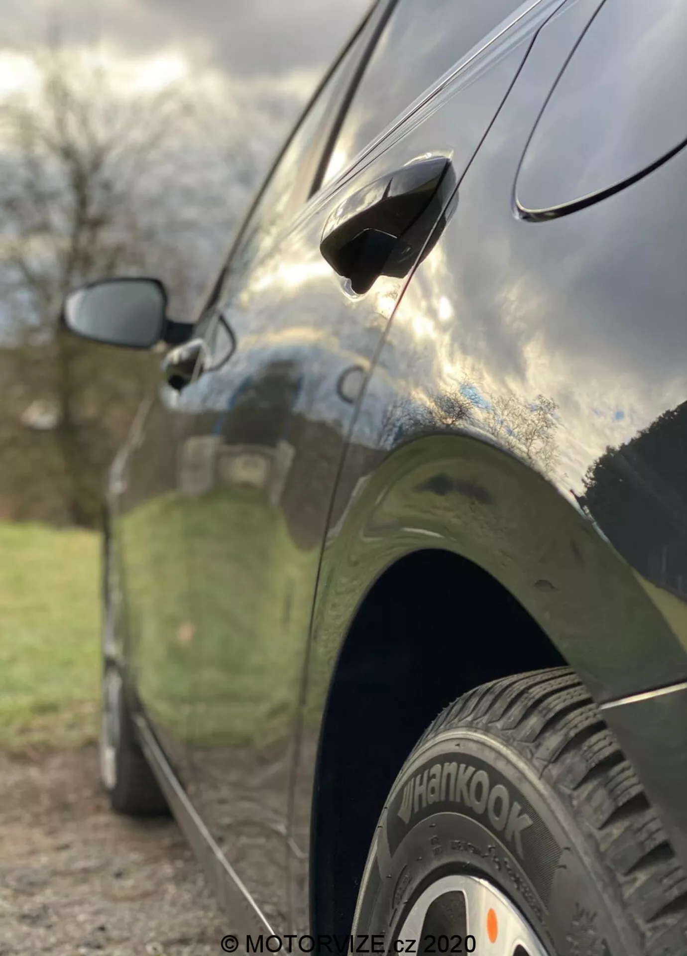 Vista laterale ravvicinata della Kia Ceed 2018, evidenziando caratteristiche come la ruota posteriore con pneumatico 'Hankook', maniglia della porta aerodinamica e specchietto laterale parzialmente sfocato. La foto è scattata da un angolo basso con una profondità di campo ridotta che mette a fuoco nitidamente la ruota posteriore e la parte inferiore della porta, mentre lo sfondo è artisticamente sfocato. L'auto ha una finitura scura e lucida che riflette il cielo e i dintorni, mostrando linee di design eleganti e una carrozzeria ben mantenuta.