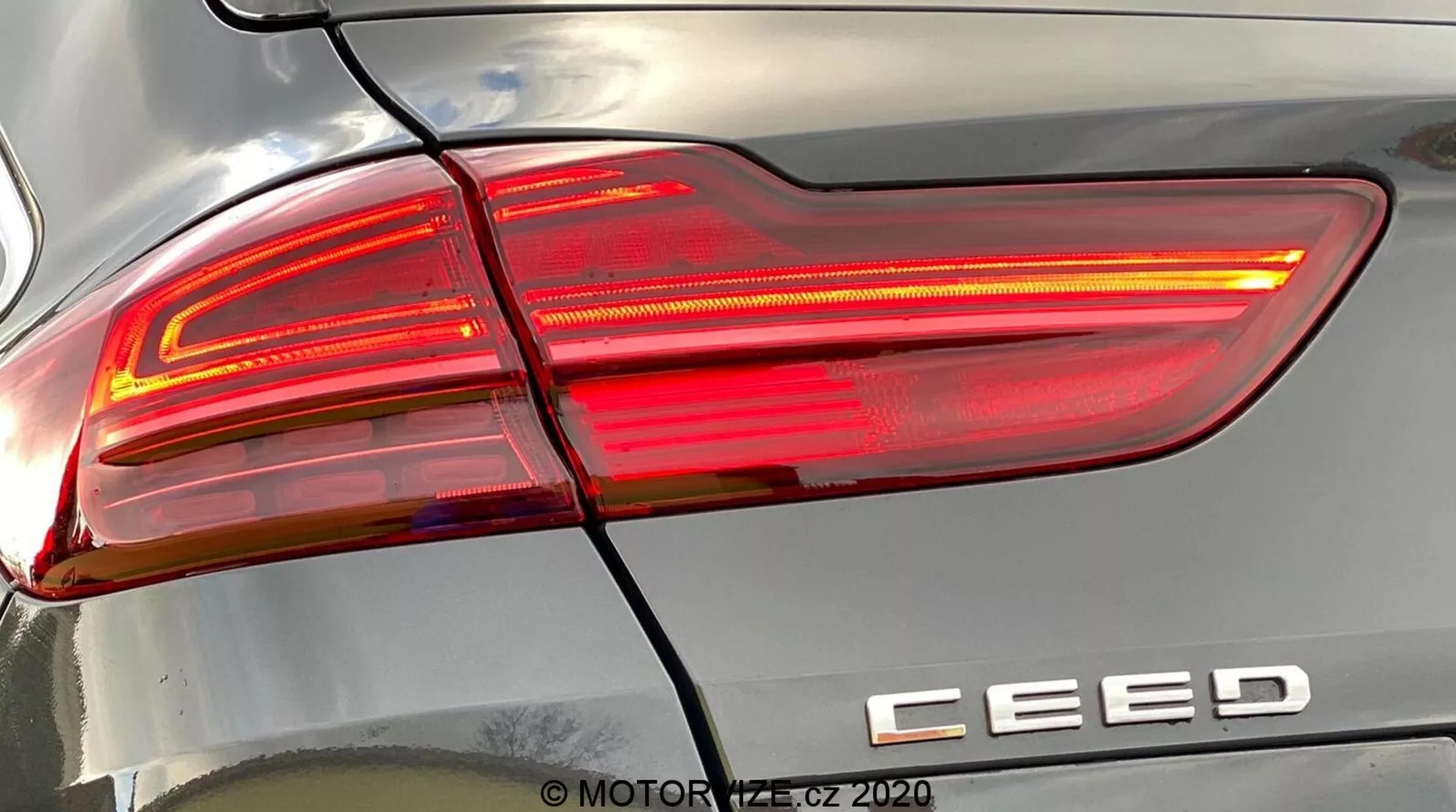 Vista dettagliata ravvicinata del gruppo fanale posteriore destro di una Kia Ceed 2018, illuminato, che mostra un design moderno con linee affilate e angolari, sezioni di lenti chiare e rosse e il badge del modello 'CEED'. Una linea orizzontale separa il fanale posteriore dal paraurti posteriore. I riflessi sulla carrozzeria evidenziano la curvatura della superficie. Filigrana 'MOTOR1.cz ©2020'.