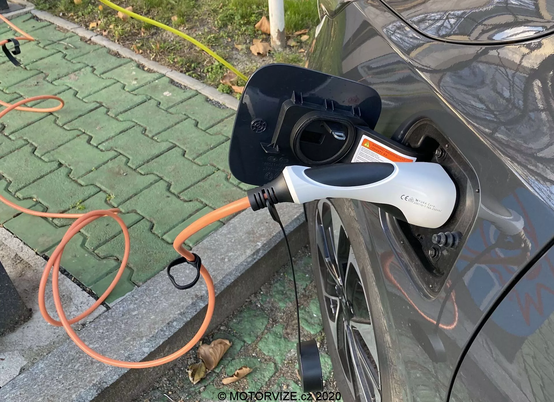 Vista laterale ravvicinata di un veicolo elettrico (EV) grigio in fase di ricarica, evidenziando la presa di ricarica aperta e un connettore EVSE arancione collegato all'auto. Il moderno connettore di ricarica bianco e nero dispone di una luce indicatrice LED. L'immagine si concentra sull'interazione di ricarica, situata in un'area pavimentata all'aperto con foglie sparse.