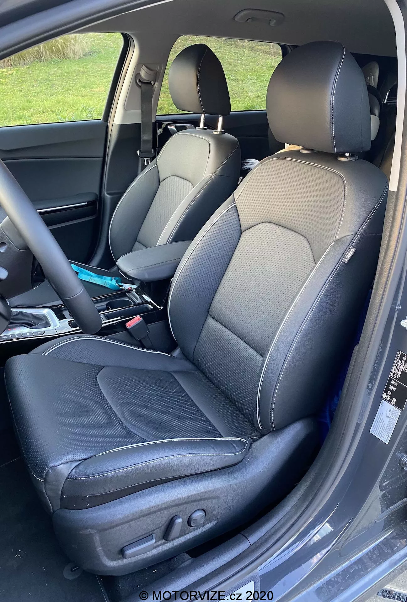 Primo piano dell'interno di una Kia Ceed 2018, che mostra i sedili del conducente e del passeggero rivestiti in pelle bicolore con cuciture a contrasto, design a tipo secchiello con supporti laterali e regolazioni elettriche per il controllo dello schienale e della posizione del sedile del conducente. Leggere pieghe indicano utilizzo e vari oggetti sono visibili sul sedile del passeggero.