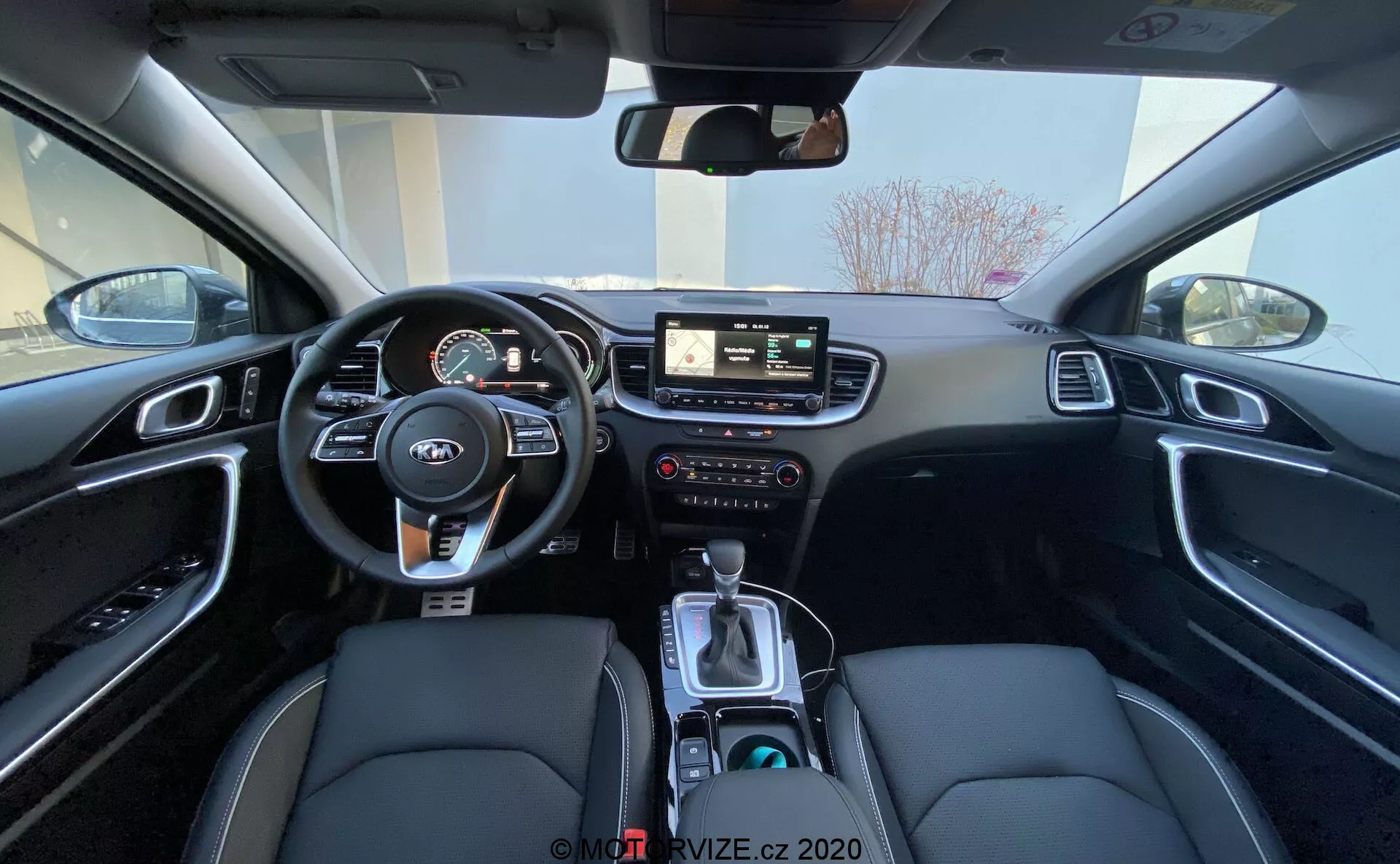 Vista grandangolare degli interni della Kia Ceed 2018 dal sedile posteriore, che mostra l'area del conducente, incluso il volante con comandi montati, il quadro strumenti con indicatori digitali e analogici, il sistema di infotainment centrale con schermo tattile, comandi manuali per infotainment e clima, e console centrale con leva del cambio, selettore della modalità di guida, portabicchieri e un interno nero con sedili in pelle sintetica con cuciture bianche e finiture in alluminio spazzolato.