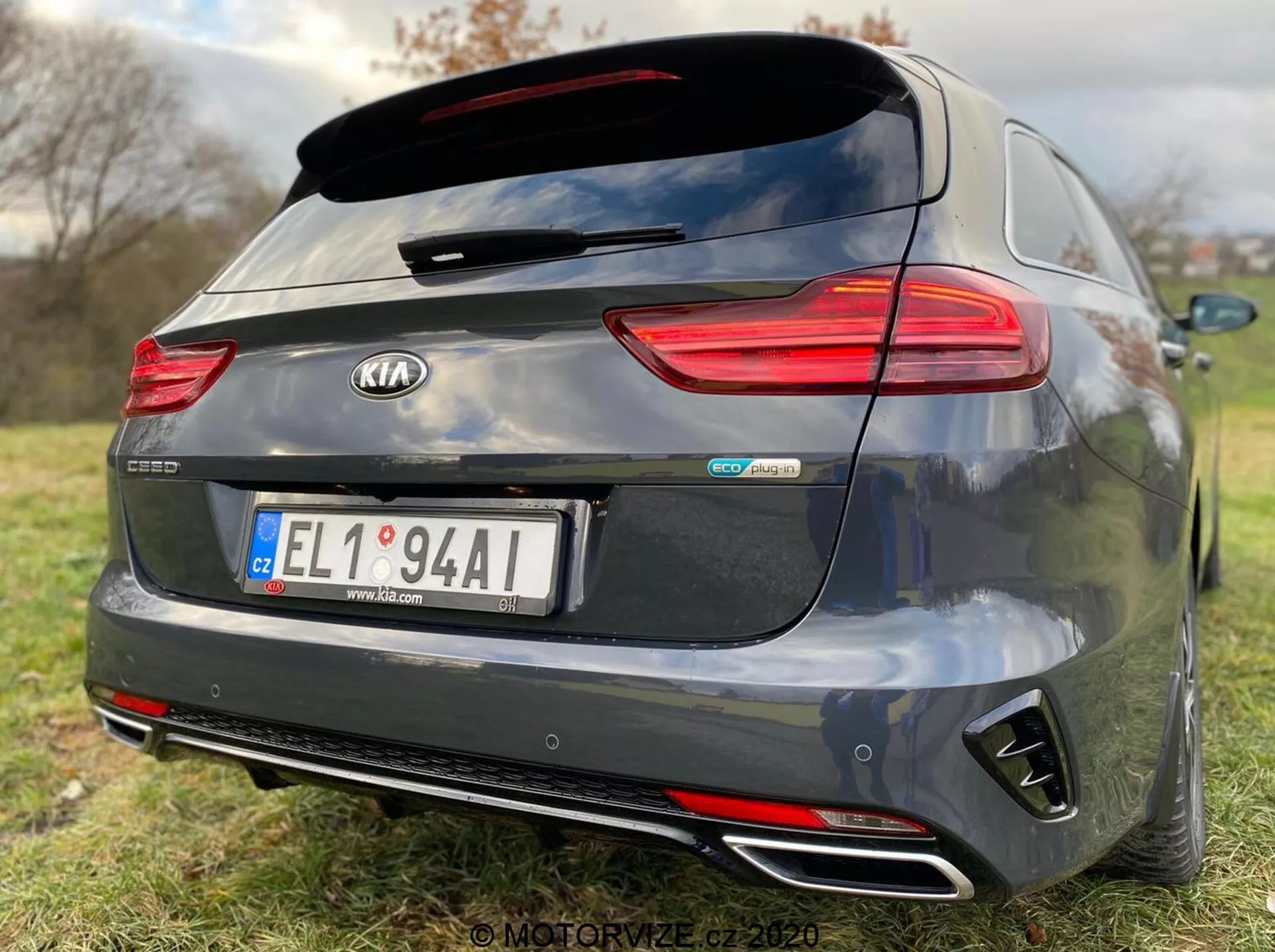 Vista posteriore di una Kia Ceed Proceed 2018 grigio scuro, con caratteristiche dettagliate come luci posteriori a LED orizzontali, spoiler posteriore montato sul tetto, doppi terminali di scarico cromati, paraurti posteriore a diffusore nero e badge centrali inclusi 'Kia' e 'GT Line.' La telecamera posteriore sopra la targa centrale suggerisce un'assistenza al parcheggio, mentre la scarsa profondità di campo enfatizza il marchio e gli identificatori del modello dell'auto su uno sfondo erboso.