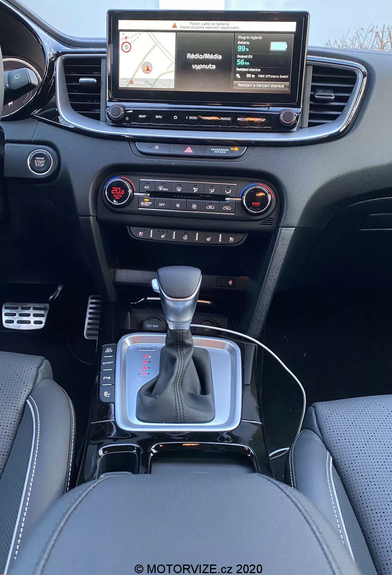 Vista interna di una Kia Ceed 2018 dalla prospettiva del sedile posteriore, mostrando un moderno cruscotto con sistema di infotainment touchscreen che mostra una mappa di navigazione, pulsanti fisici e manopole per il controllo audio e climatizzazione, bocchetta d'aria centrale, leva del cambio in posizione neutrale con accento argento e cuffia in pelle nera, piccolo spazio di stivaggio vicino alla leva del cambio possibilmente per presa 12V o porte USB, sedili in pelle con cuciture dettagliate.