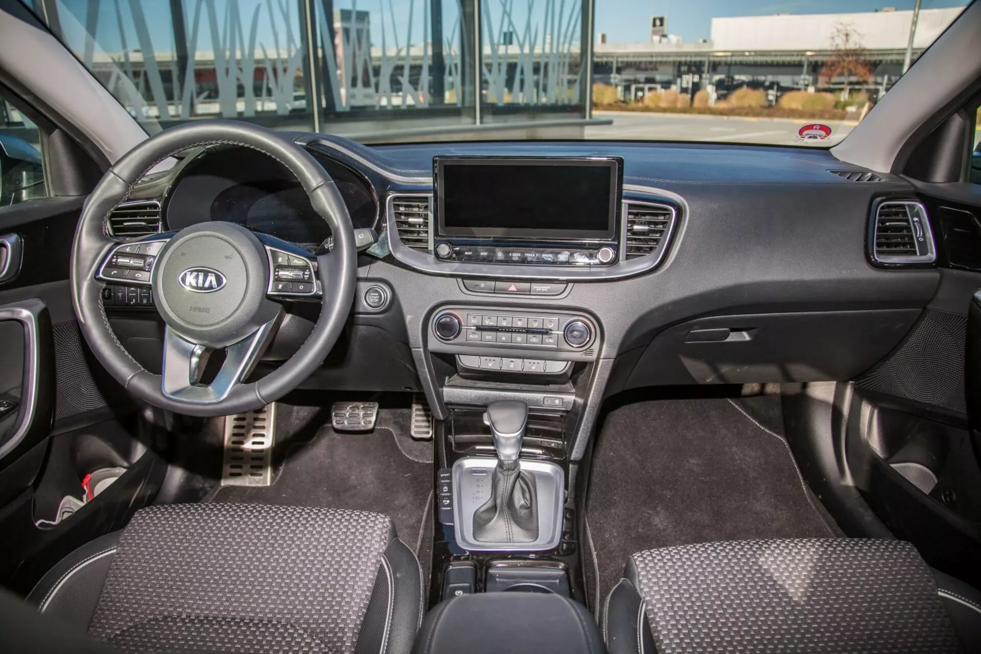 Vista interna della Kia Cee'd 2009 che mostra l'intera cabina anteriore inclusa la ruota multifunzione con logo Kia, cluster degli strumenti dietro, leva del cambio in posizione di parcheggio sulla console centrale, pulsanti per il controllo del clima, schermo del sistema di infotainment, sedili con rivestimento in tessuto e controlli porte guidatore e passeggero alla luce naturale diurna