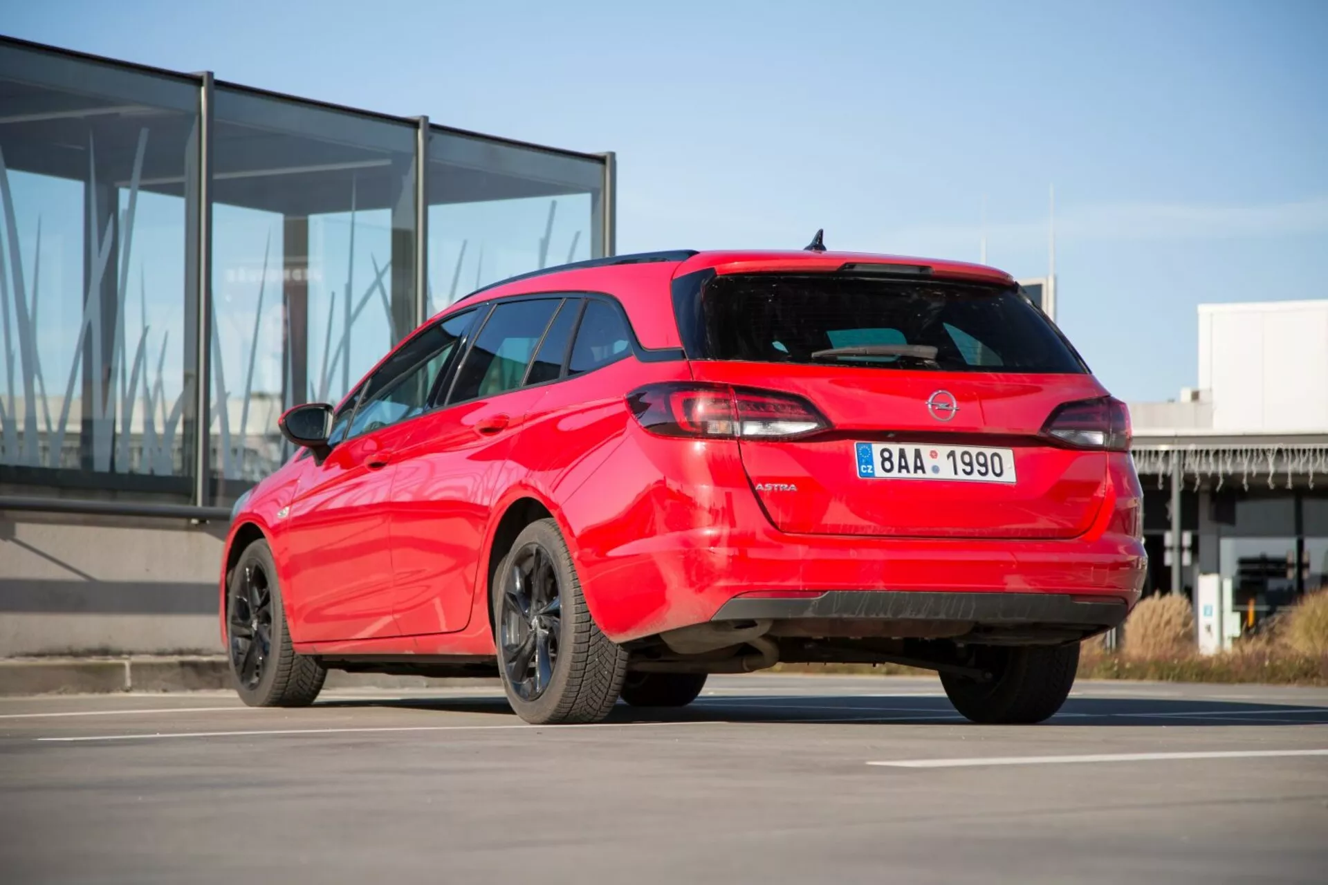Hatchback Kia Ceed del 2009 in rosso che mostra la sua vista posteriore sportiva da tre quarti su cemento con sfondo di un edificio moderno con facciata in vetro, evidenziando un design distintivo dei fanali posteriori, spoiler sul tetto, antenna a pinna di squalo, cerchi in lega e possibili sensori di parcheggio.