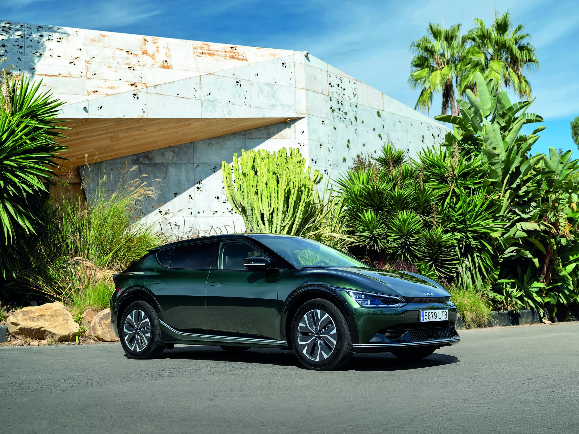 Kia EV6 2021 catturata da un angolo basso che mette in risalto il suo design elegante e l'atteggiamento sportivo con una vista frontale laterale di tre quarti che mostra fari LED, corpo aerodinamico, ruote in lega, indicatori di direzione integrati sugli specchietti laterali, parcheggiata all'aperto sotto una luce diurna brillante con un edificio moderno e vegetazione lussureggiante sullo sfondo