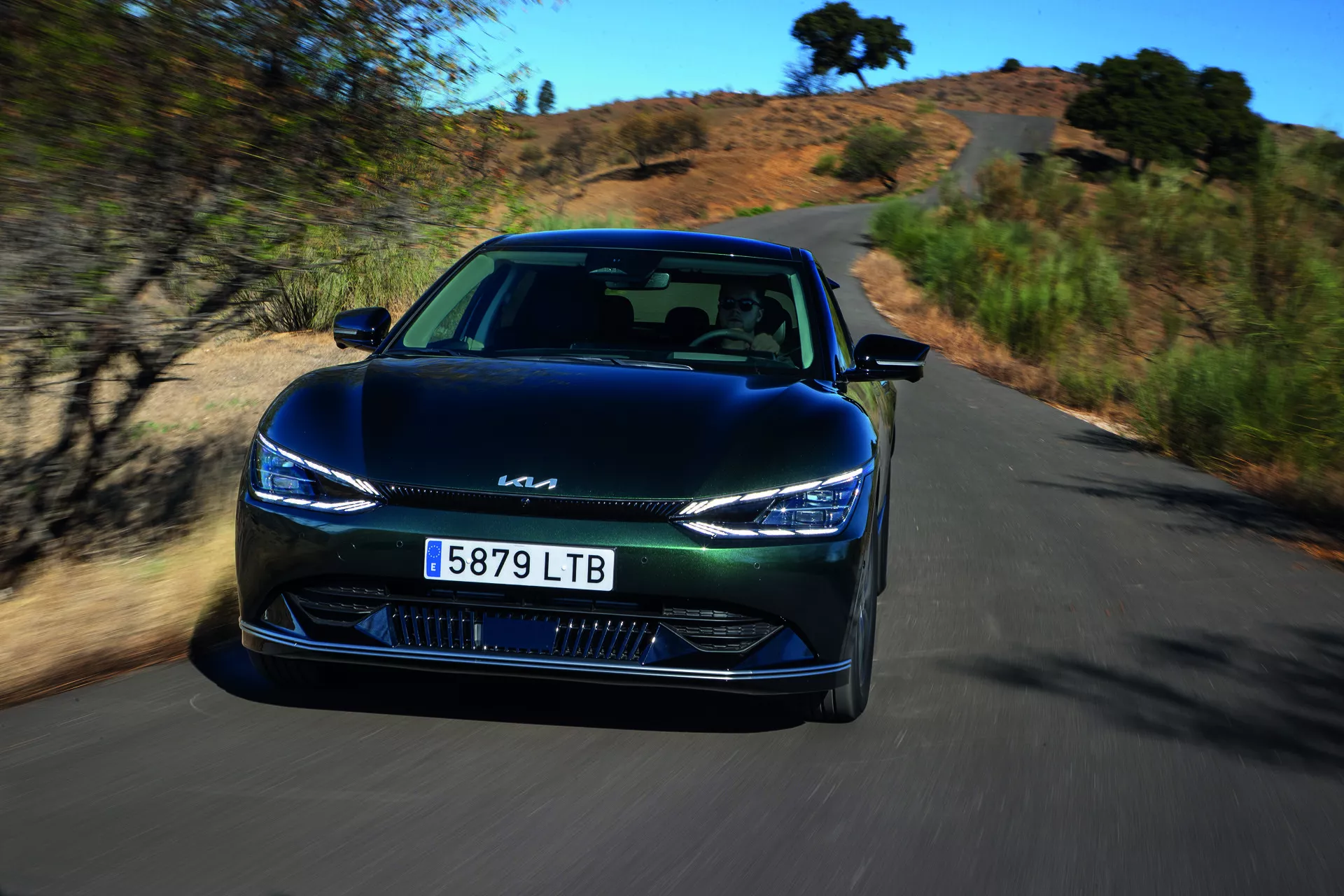 Kia EV6 2021 in movimento dinamico, vista angolare frontale che evidenzia fari LED eleganti, design della griglia chiusa e tecnologia avanzata di assistenza al guidatore su una strada panoramica naturale