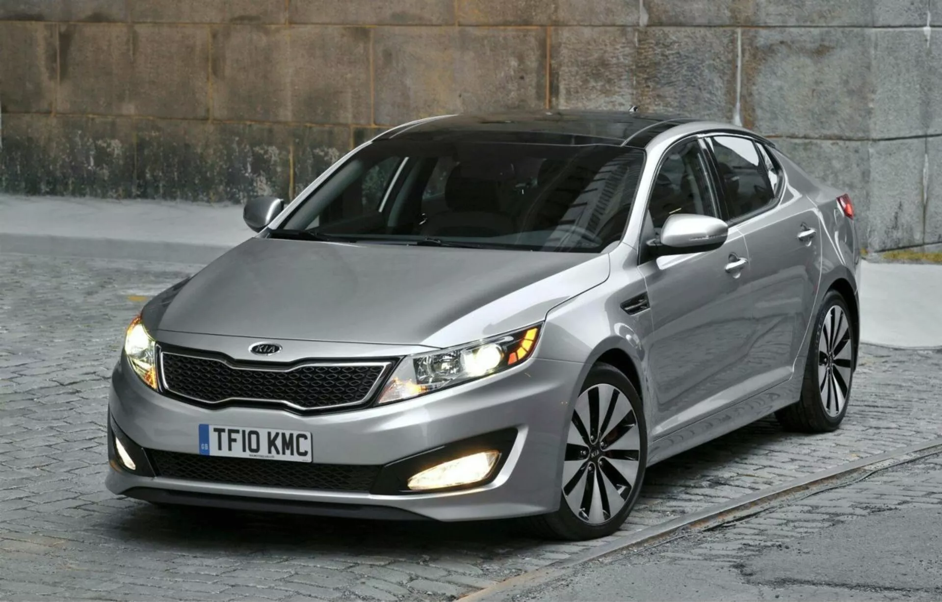Kia Optima Limousine 2012 parcheggiata su strada ciottolata mostrando il suo quarto anteriore sinistro inclusi la griglia con accenti cromati, fari a lente chiara, luci di nebbia, vernice metallica, ruote in lega a più razze e specchietti con indicatori di direzione integrati, sottolineando il suo design aerodinamico e le caratteristiche moderne da berlina in un contesto urbano