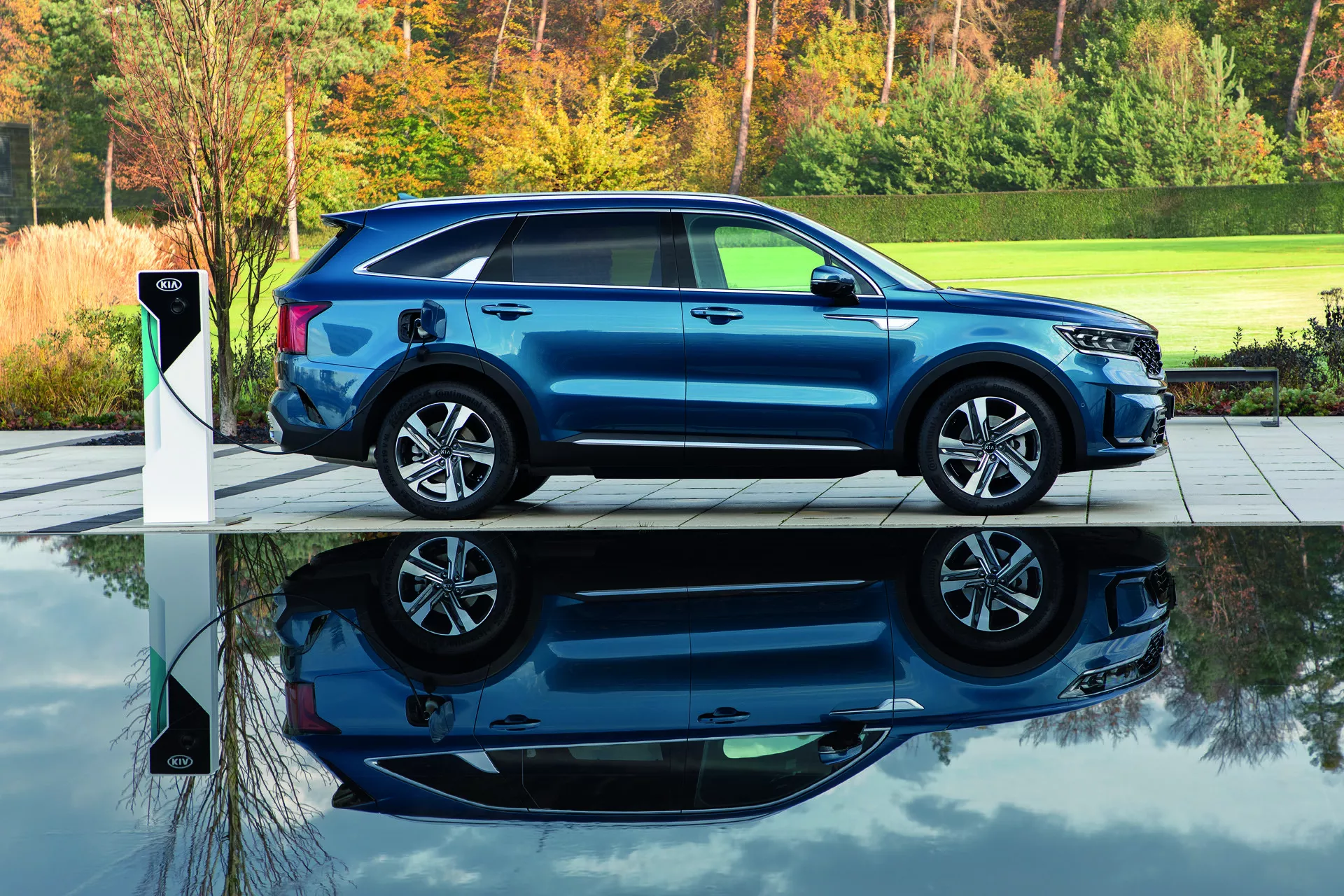 SUV blu moderno Kia Sorento MQ4 2020 parcheggiato su pavimentazione grigia accanto a stazione di ricarica per veicoli elettrici, mostrando profilo laterale con vista frontale parziale, evidenziato da luce naturale mattutina o serale con sfondo di fogliame verde, sottolineando la capacità di ricarica elettrica, design aerodinamico e ruote in lega.