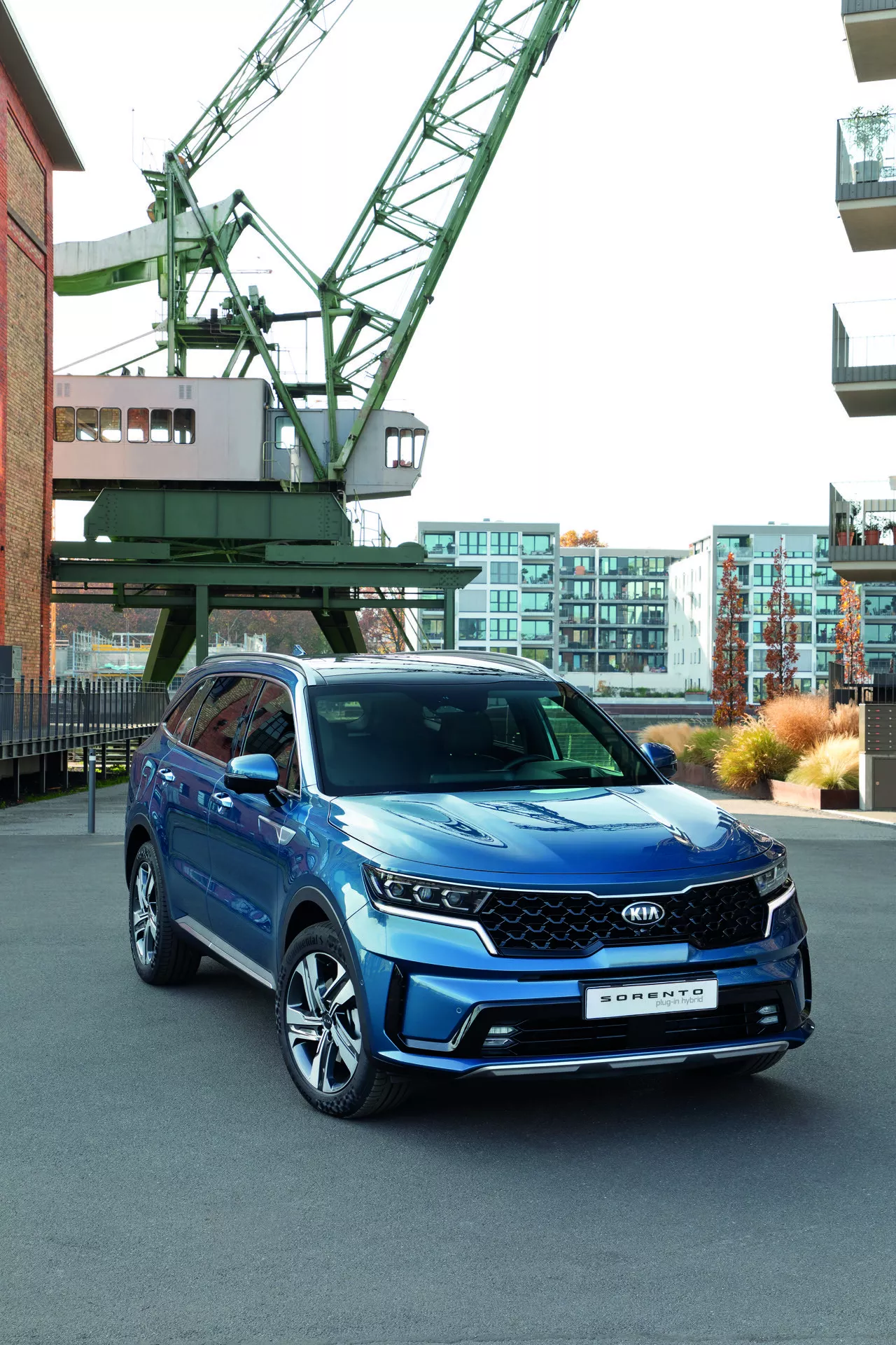 Kia Sorento MQ4 del 2020 in blu esposta in un contesto urbano con gru industriale e moderni edifici sullo sfondo, mettendo in evidenza fari LED, griglia geometrica con possibili sensori ADAS, ruote in lega a più razze e indicatori di direzione integrati negli specchietti laterali, sottolineando le sue caratteristiche tecnologiche e l'aspetto sportivo.