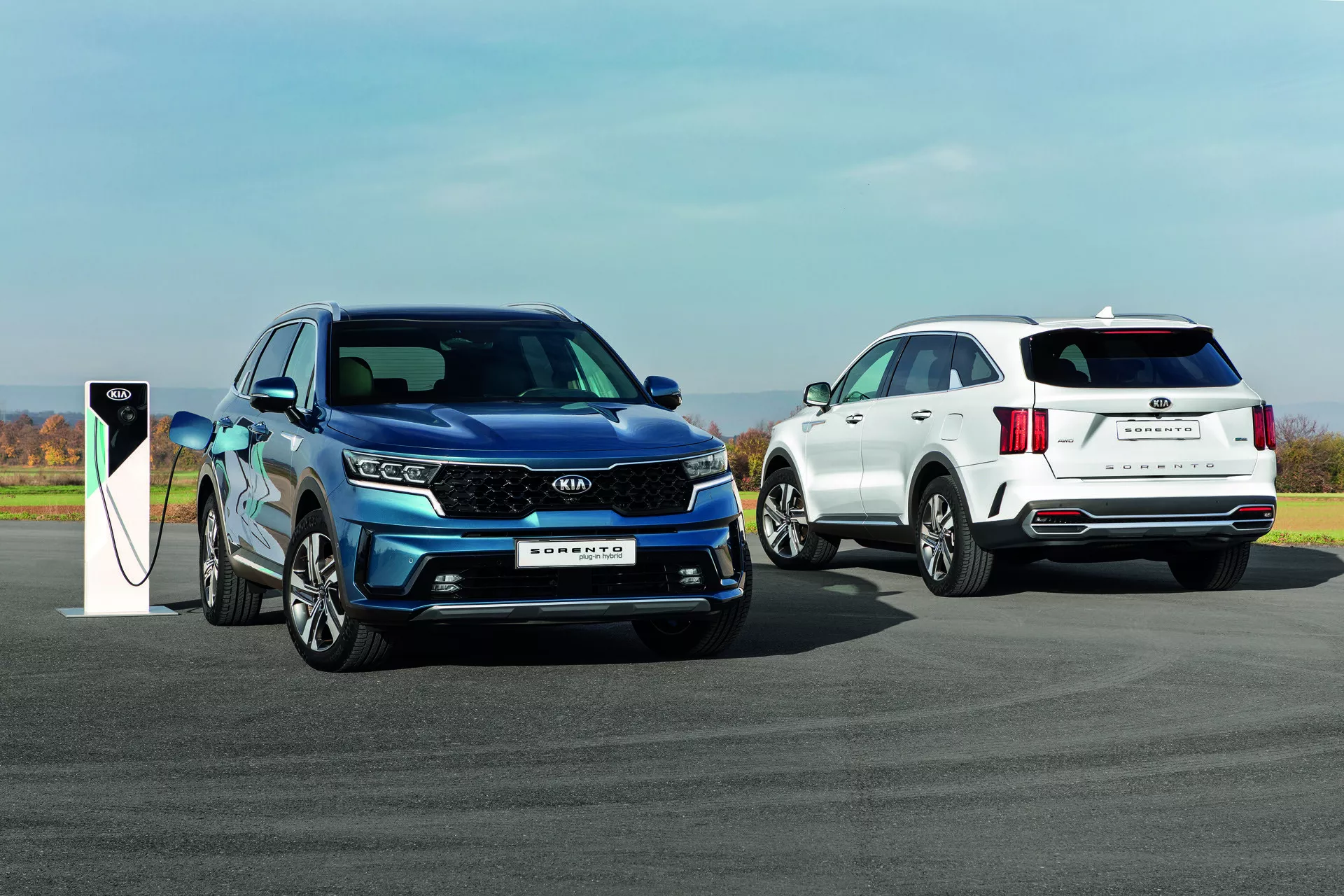 Kia Sorento MQ4 del 2020 in blu, modello elettrico o ibrido plug-in in carica, e un SUV bianco sullo sfondo su una superficie esterna pavimentata sotto un cielo sereno