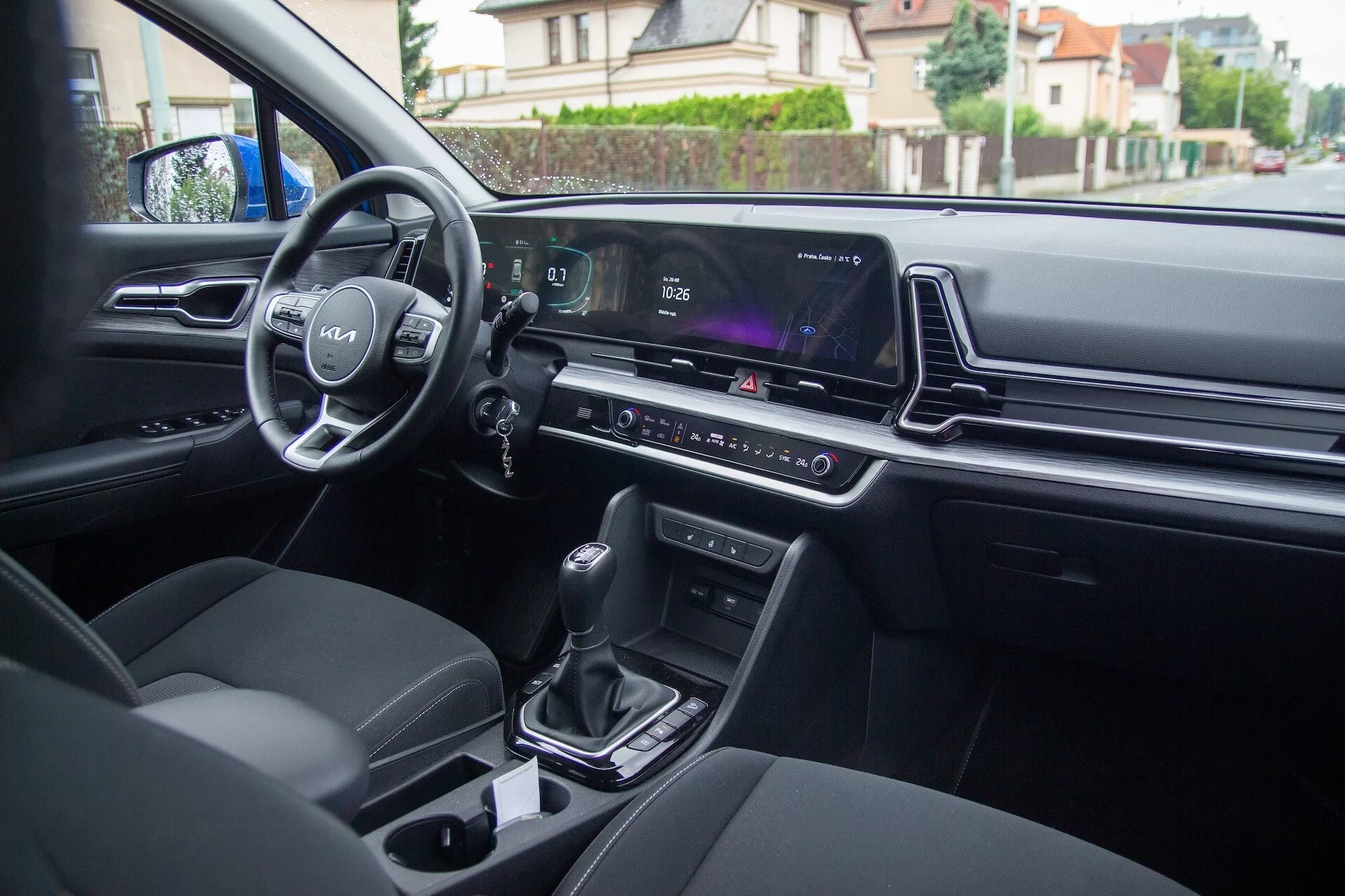 Vista interna della Kia Sportage 2022 dal sedile posteriore che mostra i controlli sulla porta del conducente, volante con logo e controlli montati, quadro strumenti digitale, schermo infotainment, controllo clima, leva del cambio automatica, portabicchieri e sedili imbottiti moderni