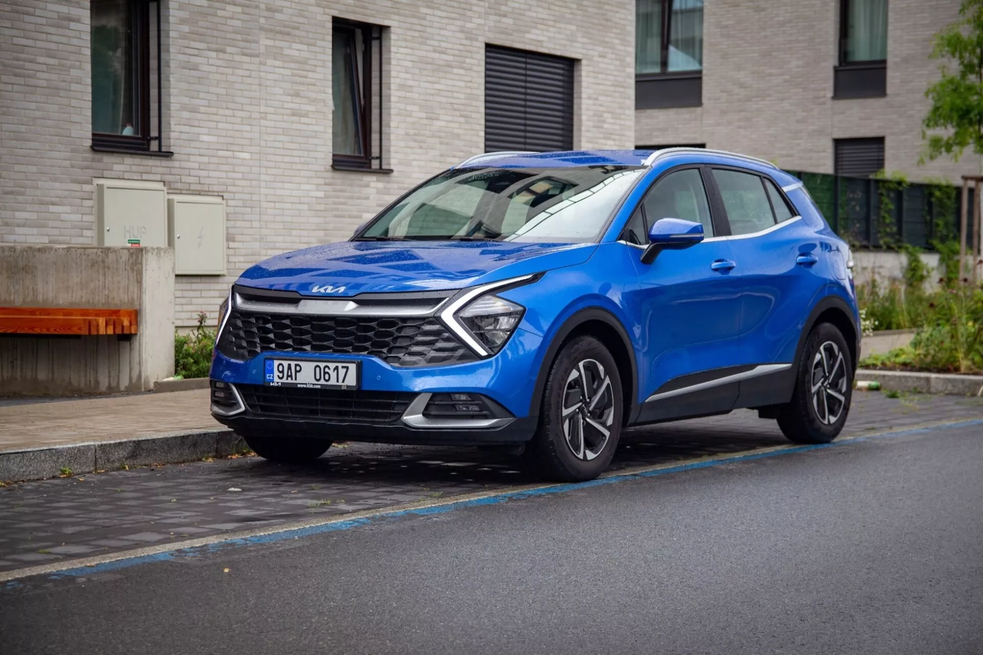 SUV crossover compatto Kia Sportage 2022 di colore blu, parcheggiato su una strada bagnata mostrando la sua visuale dinamica frontale a tre quarti con luci diurne a LED, cofano scolpito e ruote in lega.