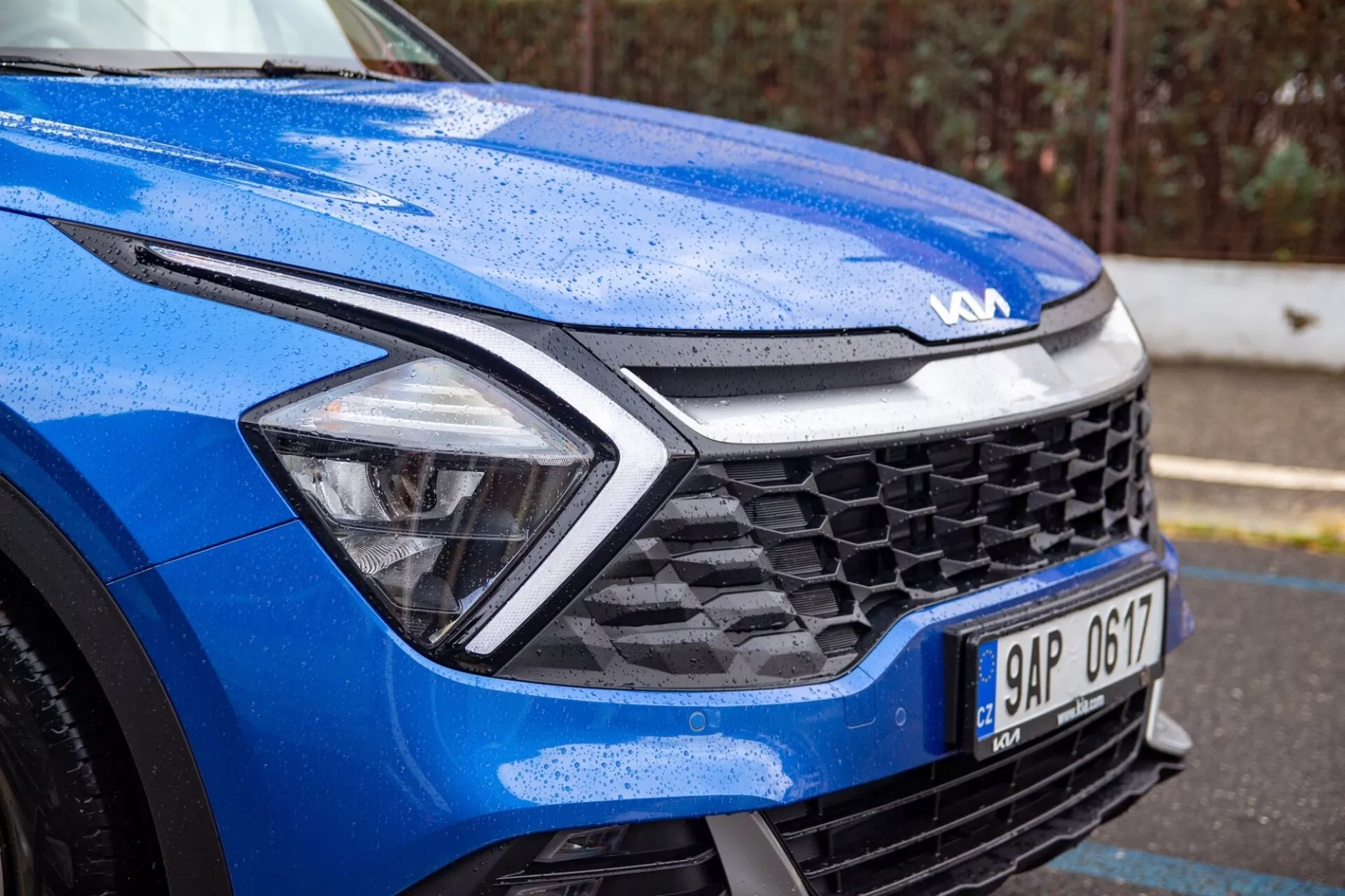Vista ravvicinata di una Kia Sportage blu 2022 concentrata sul quarto anteriore sinistro in condizioni di bagnato, evidenziando il design dinamico del cofano, fari LED, griglia a nido d'ape e tecnologia dei sensori avanzata
