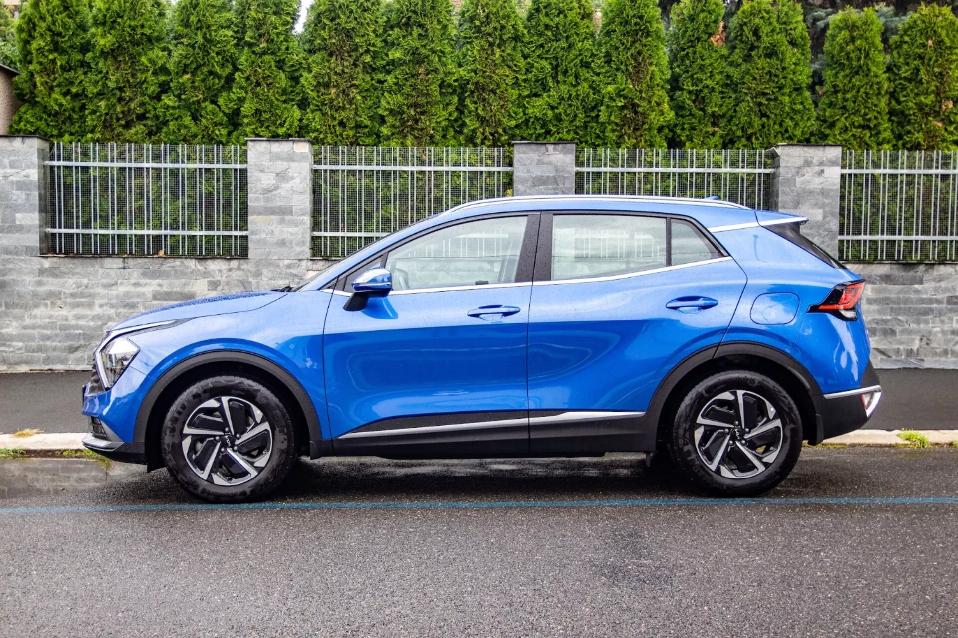 SUV crossover Kia Sportage 2022 in blu brillante, che mostra il suo design elegante e compatto con un tetto inclinato in stile coupé, cofano contornato, unità di fari LED, cerchi in lega a cinque razze e miglioramenti aerodinamici come uno spoiler posteriore, in un ambiente suburbano.