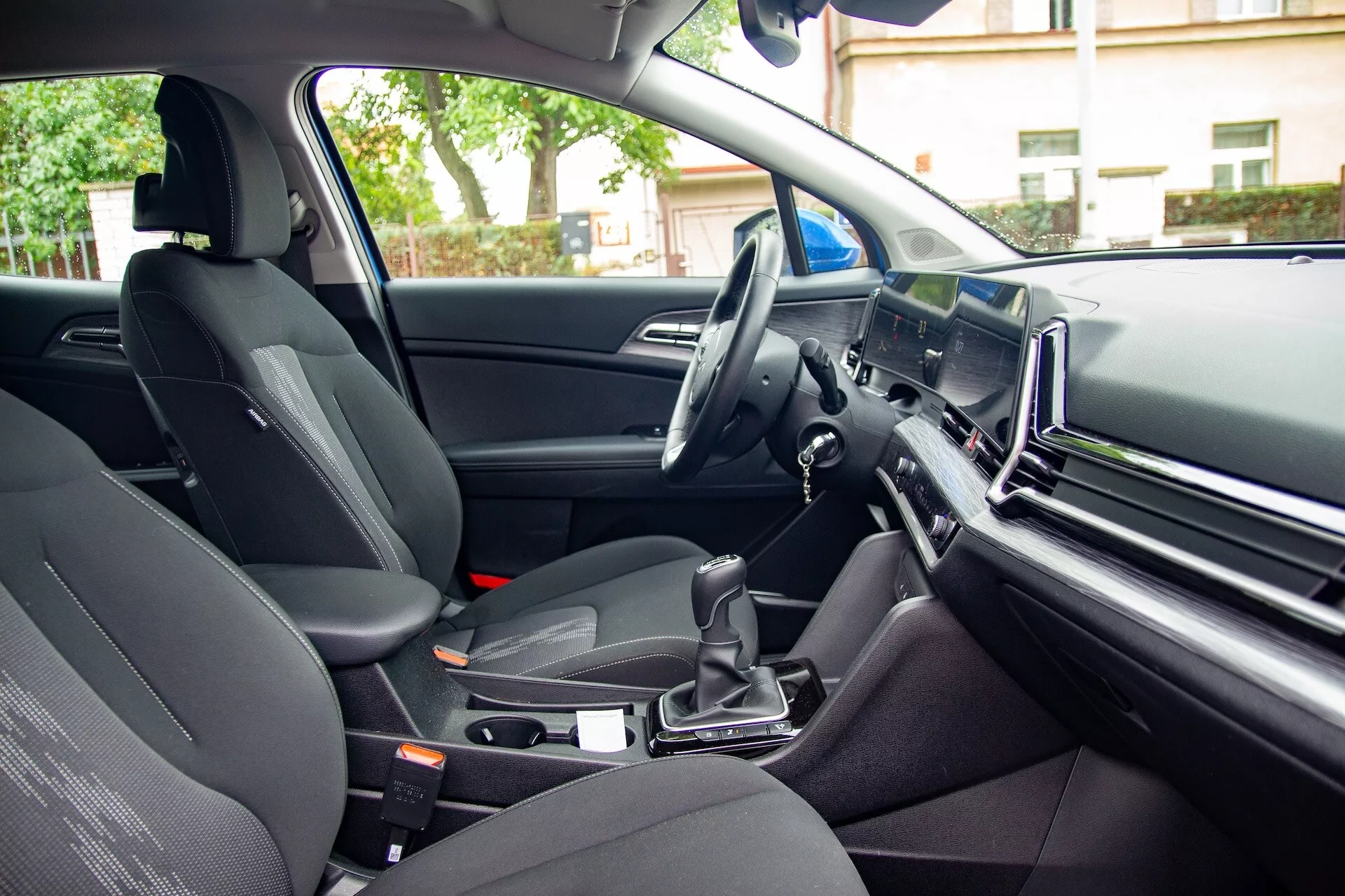 Vista interna della Kia Sportage 2022 dal punto di vista del passeggero posteriore mostrando il sedile del passeggero anteriore, la console centrale con la leva del cambio automatico, il volante a sinistra, e un grande schermo infotainment sul cruscotto