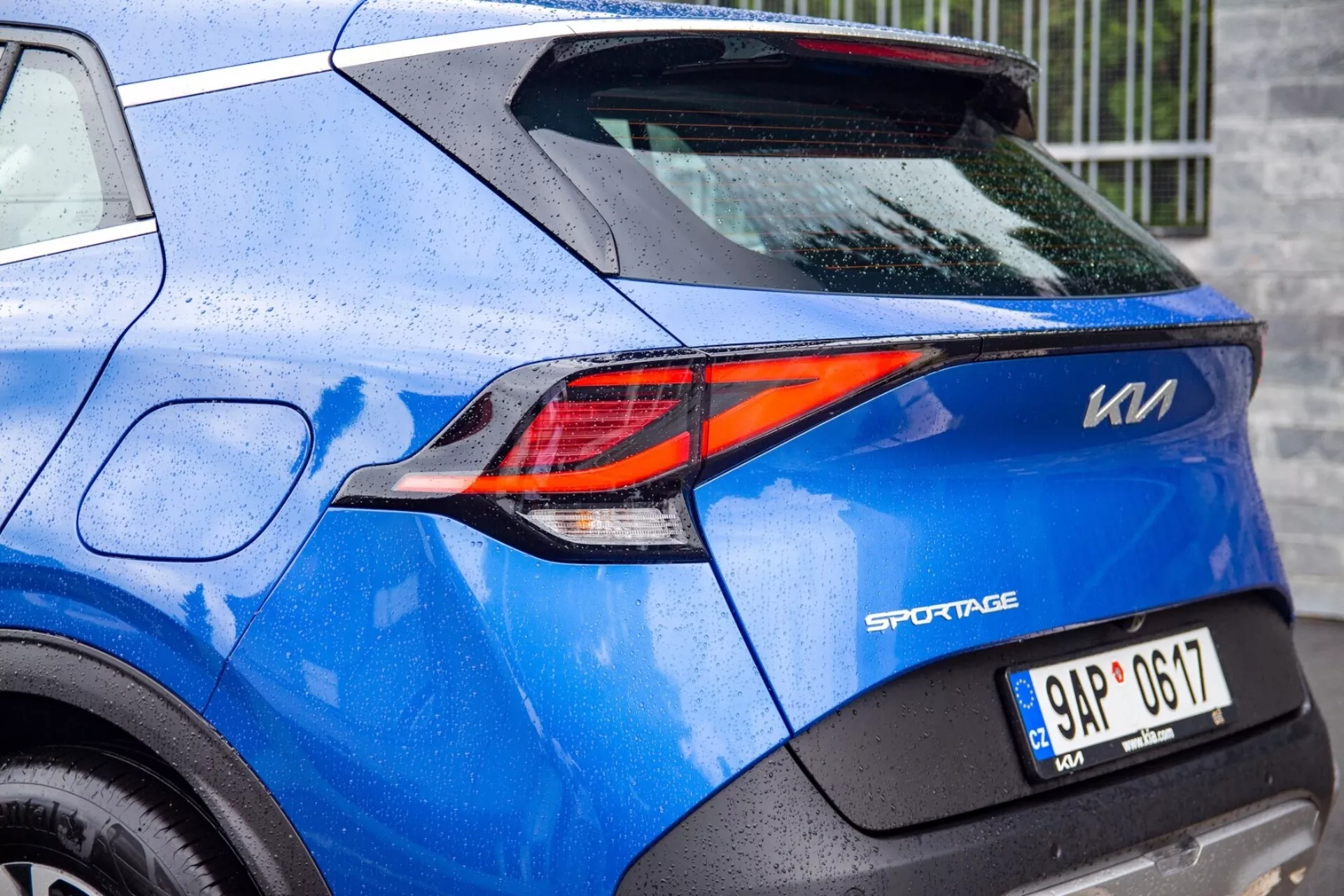 Vista parziale di un Kia Sportage blu 2022 che mette in evidenza la sofisticata luce posteriore LED a forma di Z e i dettagli del design posteriore, inclusi il distintivo del modello, la targa e la vernice punteggiata di pioggia
