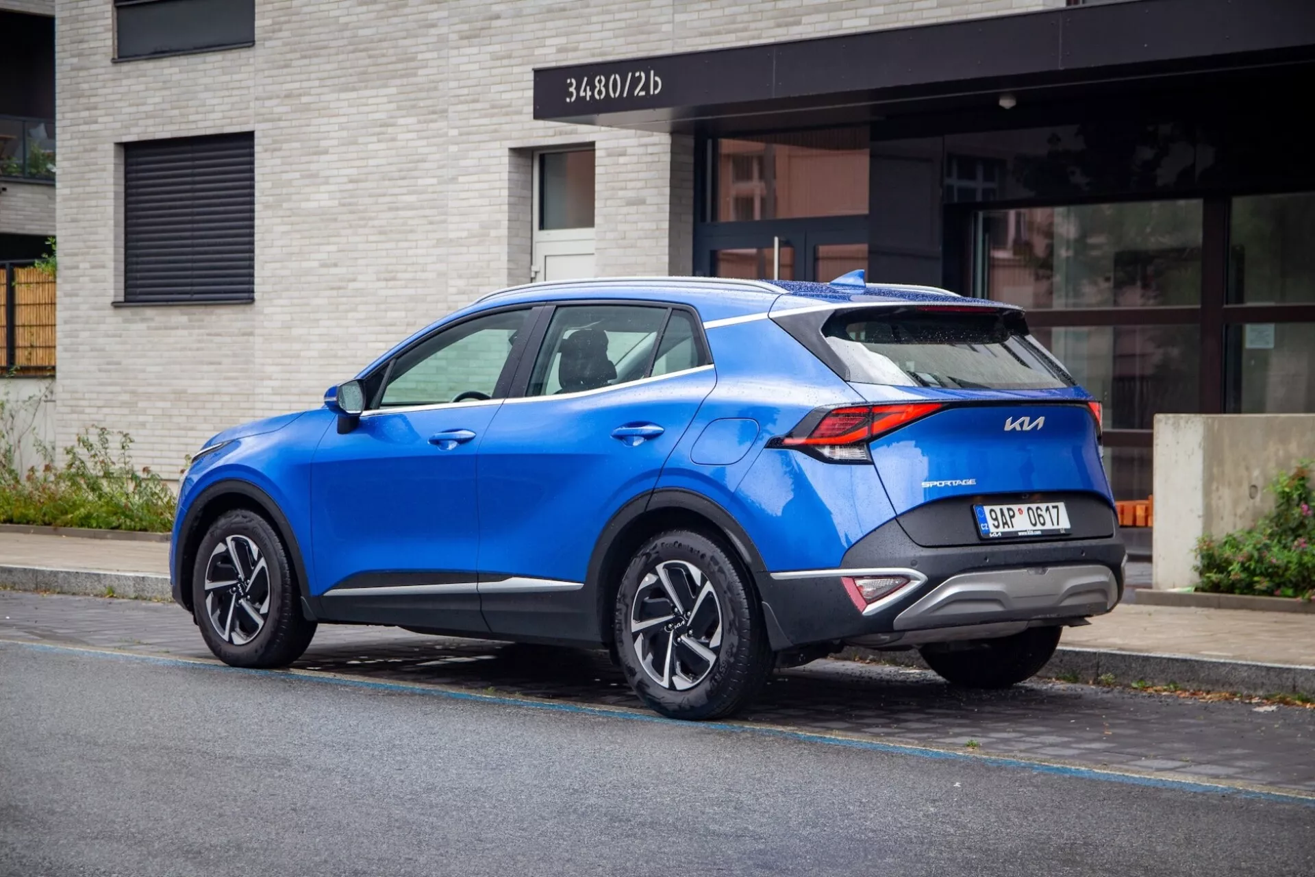 SUV crossover moderno Kia Sportage 2022 parcheggiato su una strada cittadina, mostrando vista posteriore tre quarti con fanali distintivi, spoiler sul tetto sportivo, ruote in lega a più razze, colore blu vivace e potenziali caratteristiche tecnologiche moderne.