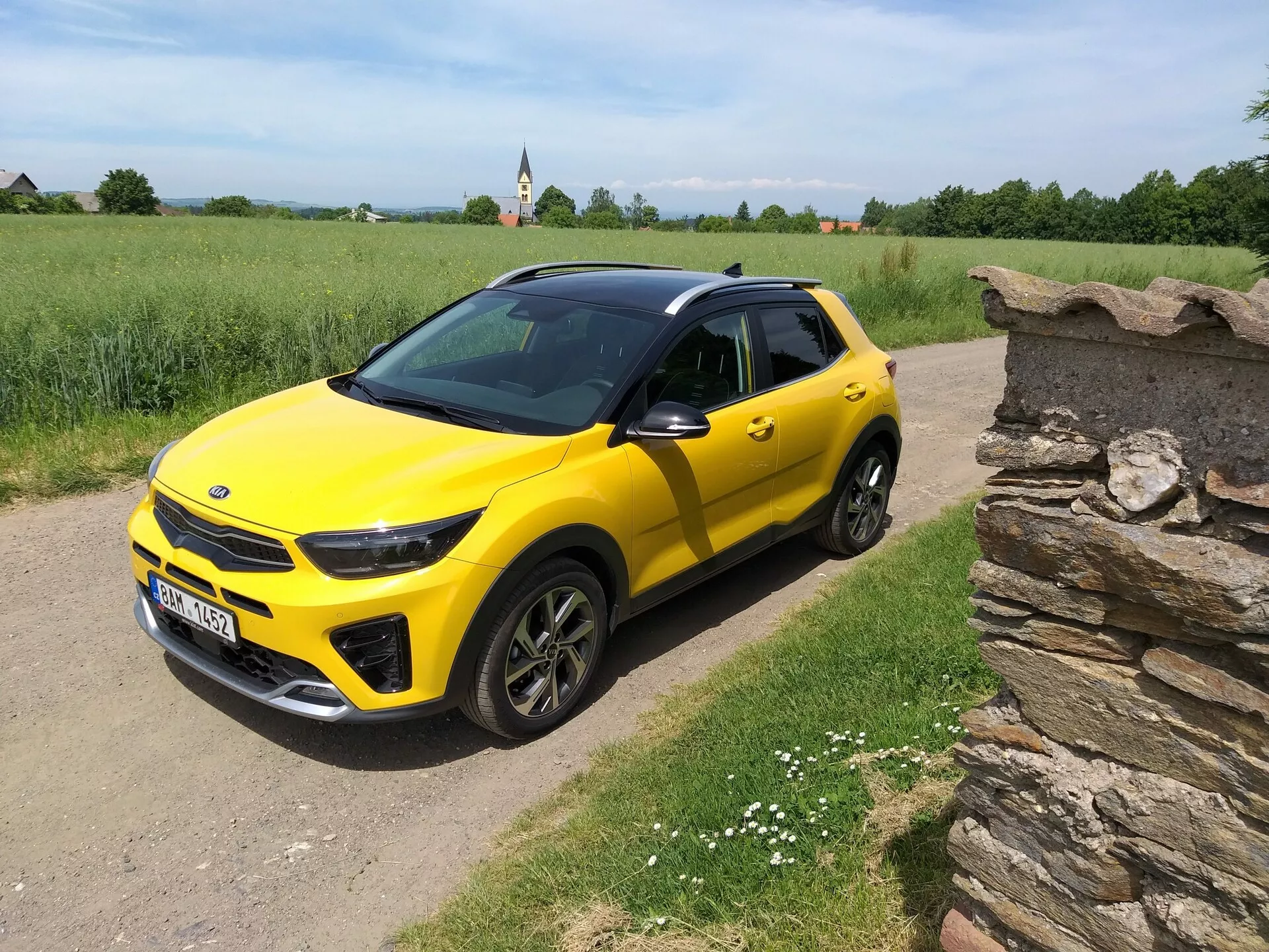 SUV compatto Kia Stonic 2017 giallo brillante con tetto nero, che mostra il suo design elegante e i contorni dinamici da un angolo frontale di tre quarti su un sentiero di campagna