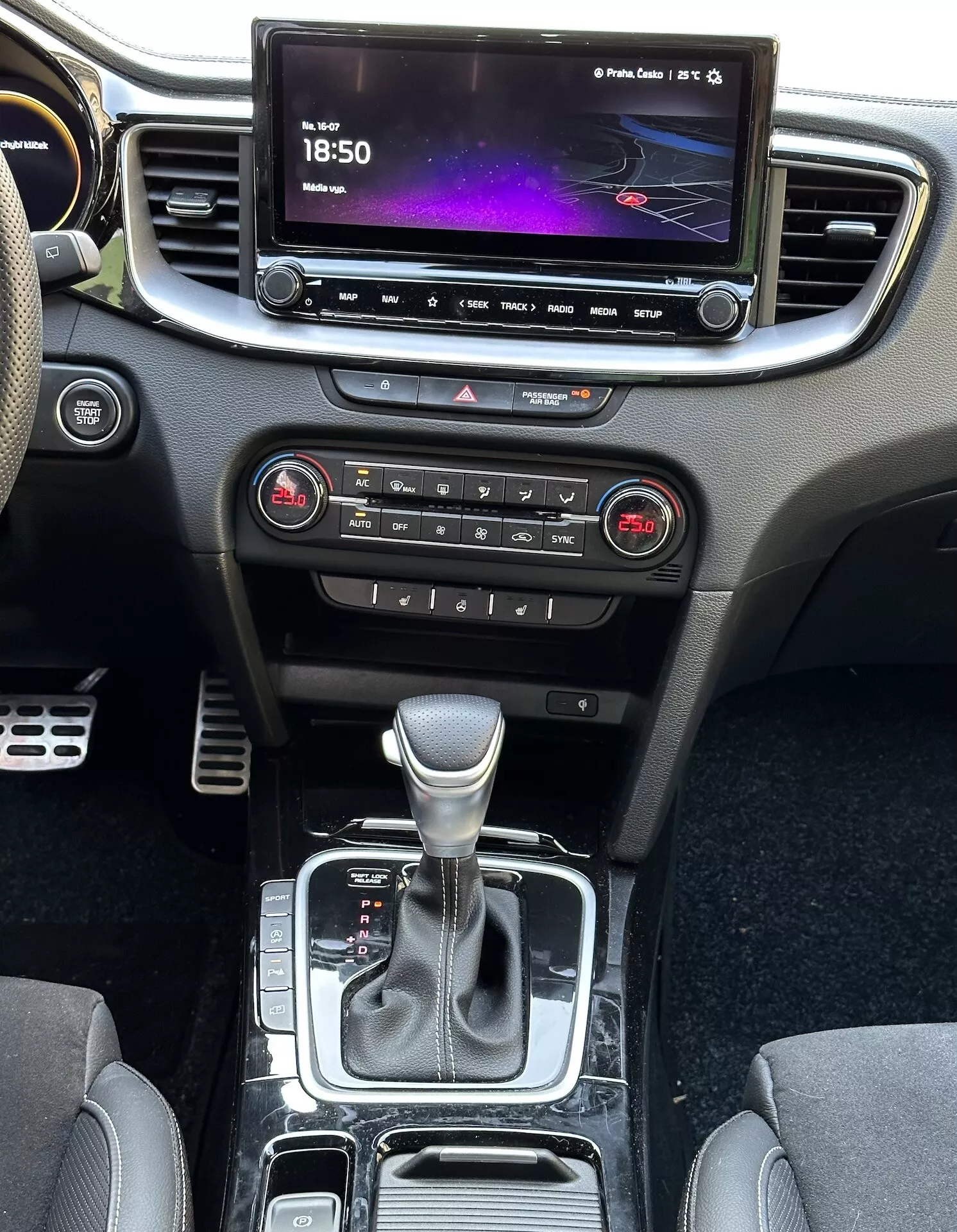 Vista interna della Kia Xceed 2022 che mostra la console centrale, il cruscotto, lo schermo dell'infotainment, i pulsanti per il controllo del clima, la leva del cambio e le aree di stoccaggio, evidenziando la tecnologia moderna e il design del veicolo.
