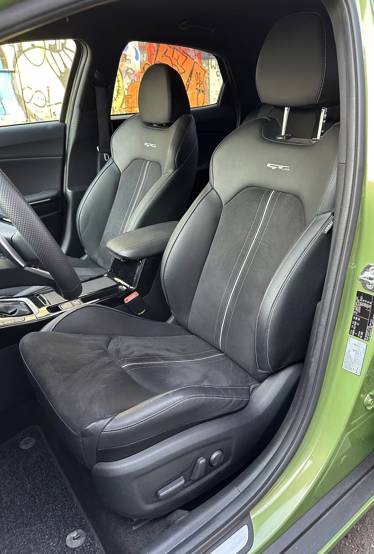 Vista interna della Kia XCeed 2022 che si concentra sul compartimento passeggeri anteriore rivestito in pelle con cuciture intricate e comandi di regolazione del sedile elettronici, da un angolo di porta aperta che mostra un accenno dell'esterno verde vibrante.