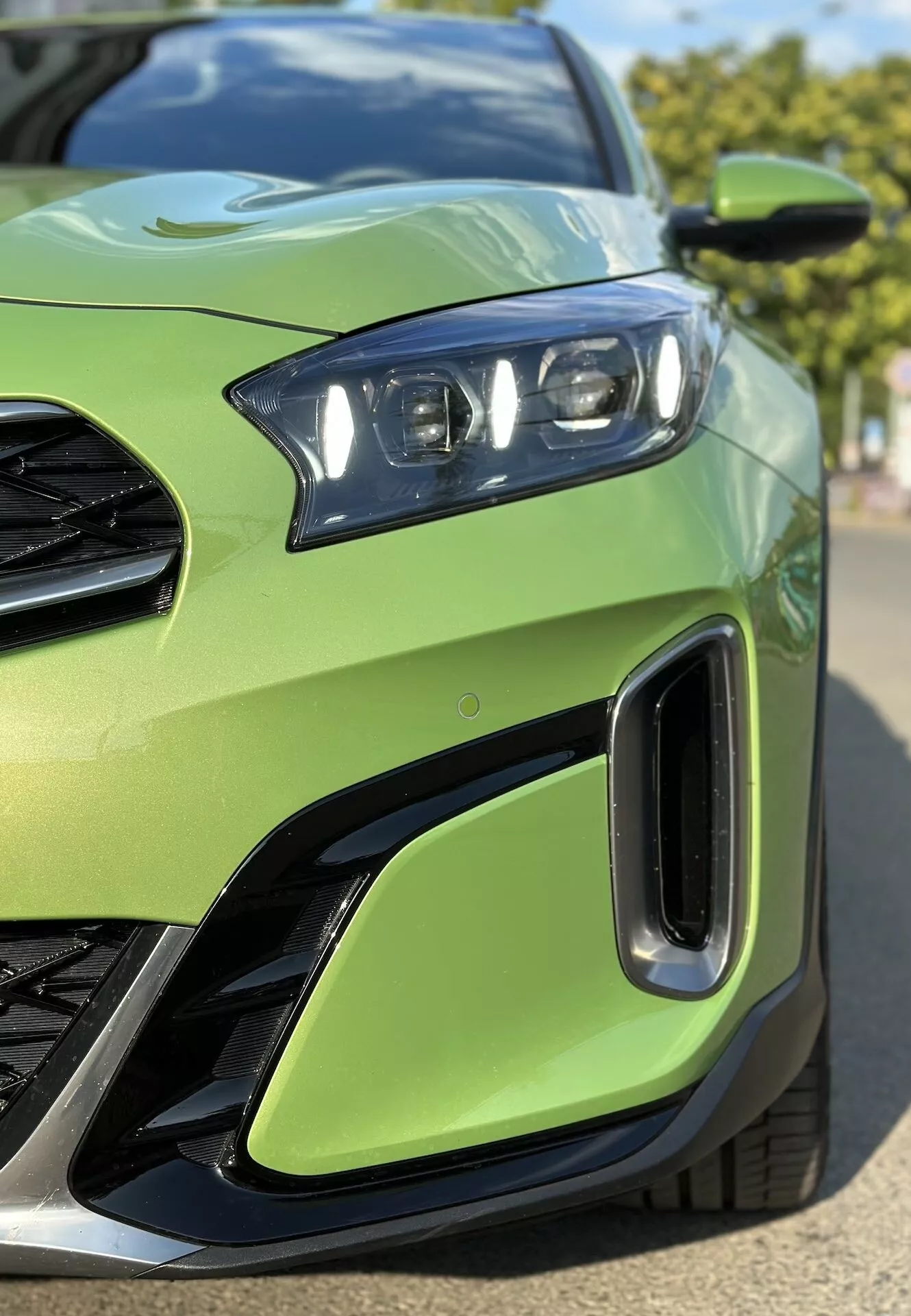 Primo piano del quarto anteriore sinistro della Kia Xceed 2022 con vista dettagliata dell'assemblaggio del faro LED, parte della griglia, prese d'aria sul paraurti anteriore, impostato su un colore della carrozzeria verde lime, sottolineando il design sportivo del veicolo e la tecnologia di illuminazione avanzata.