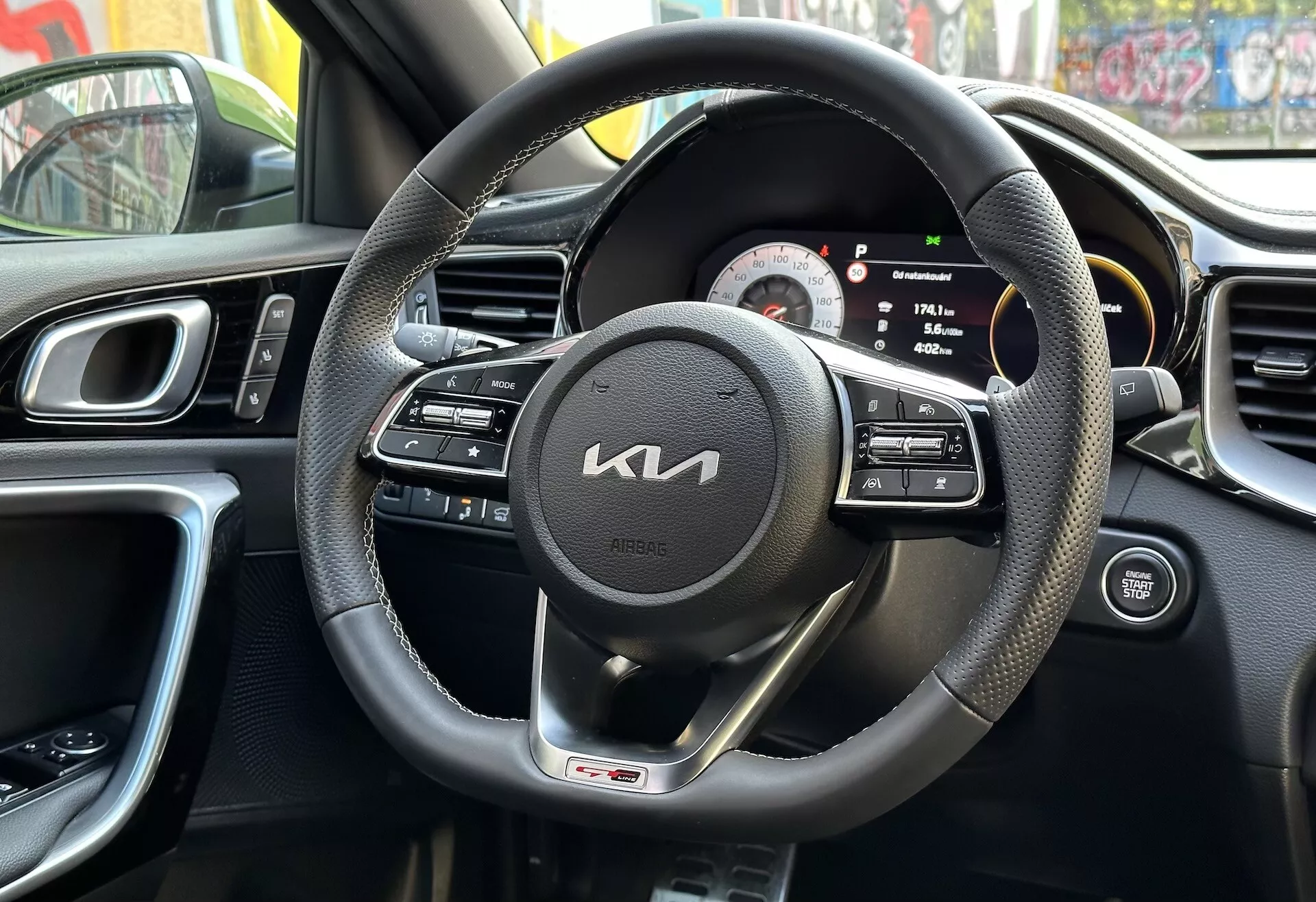 Vista interna Kia XCeed 2022 dalla prospettiva del conducente con volante avvolto in cuoio nero con controlli multifunzione e distintivo GT line, cluster strumenti digitale-analogico, parte della consola centrale con bocchette d'aria, design ergonomico del cruscotto, e pannello porta del conducente con comandi, riflettendo ambiente urbano