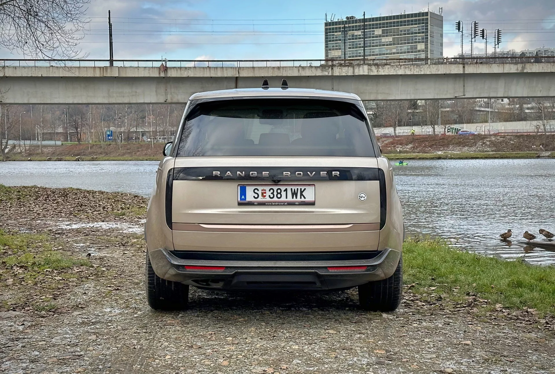 Vista posteriore di Range Rover 2021 con angolazione diretta all'altezza degli occhi, parcheggiata vicino a uno specchio d'acqua con anatre e un cavalcavia sullo sfondo, che mostra il nome del modello, luci posteriori, sensori di parcheggio e sistema di scarico doppio in beige opaco