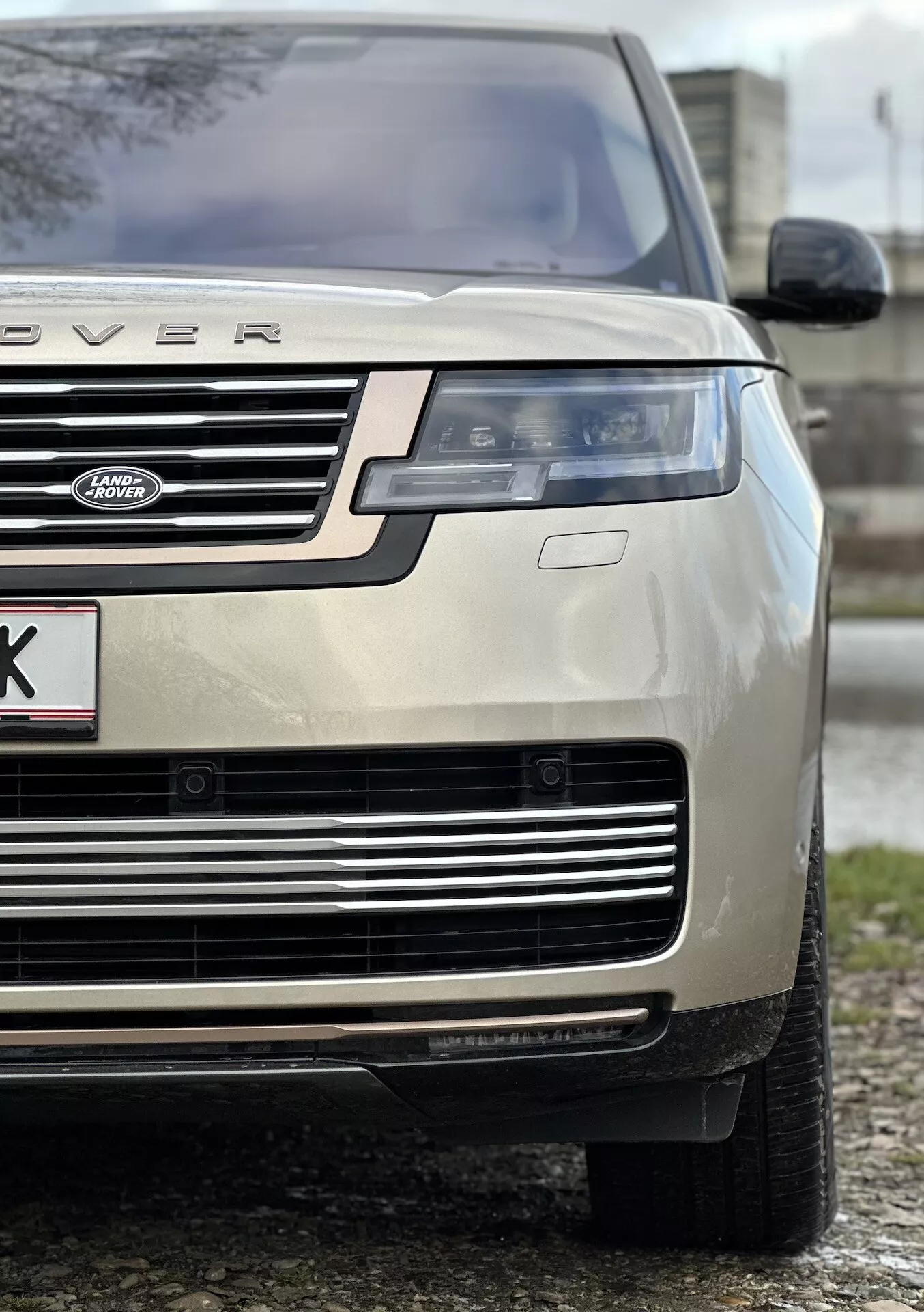 Primo piano di un Land Rover Range Rover color oro del 2021 che evidenzia il quarto anteriore sinistro, enfatizzando l'altezza e la posizione del veicolo con un focus sul cofano, la griglia, il faro LED, il paraurti e il pneumatico per tutte le stagioni, indicativo di lusso e capacità fuoristrada.