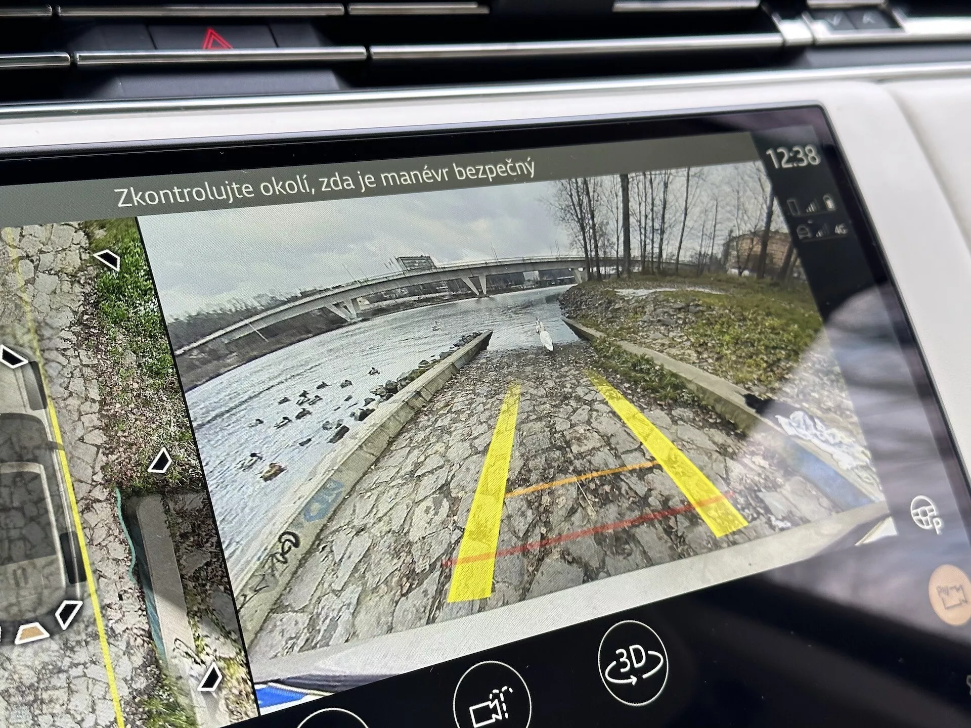 Display infotainment di Land Rover Range Rover 2021 che mostra la vista posteriore dalla telecamera di retromarcia con linee guida dinamiche in un'area parcheggio vicino a un'area d'acqua, dettagliata con vegetazione, icone di assistenza al parcheggio e testo di promemoria di sicurezza